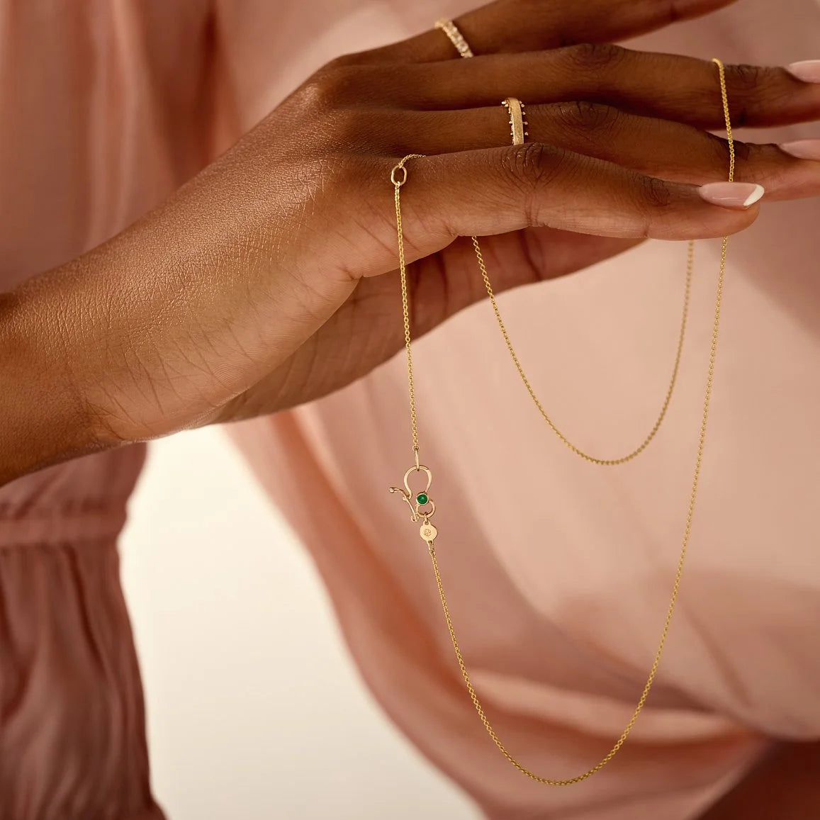 Gold necklace with a pendant held by a hand against a soft pink background