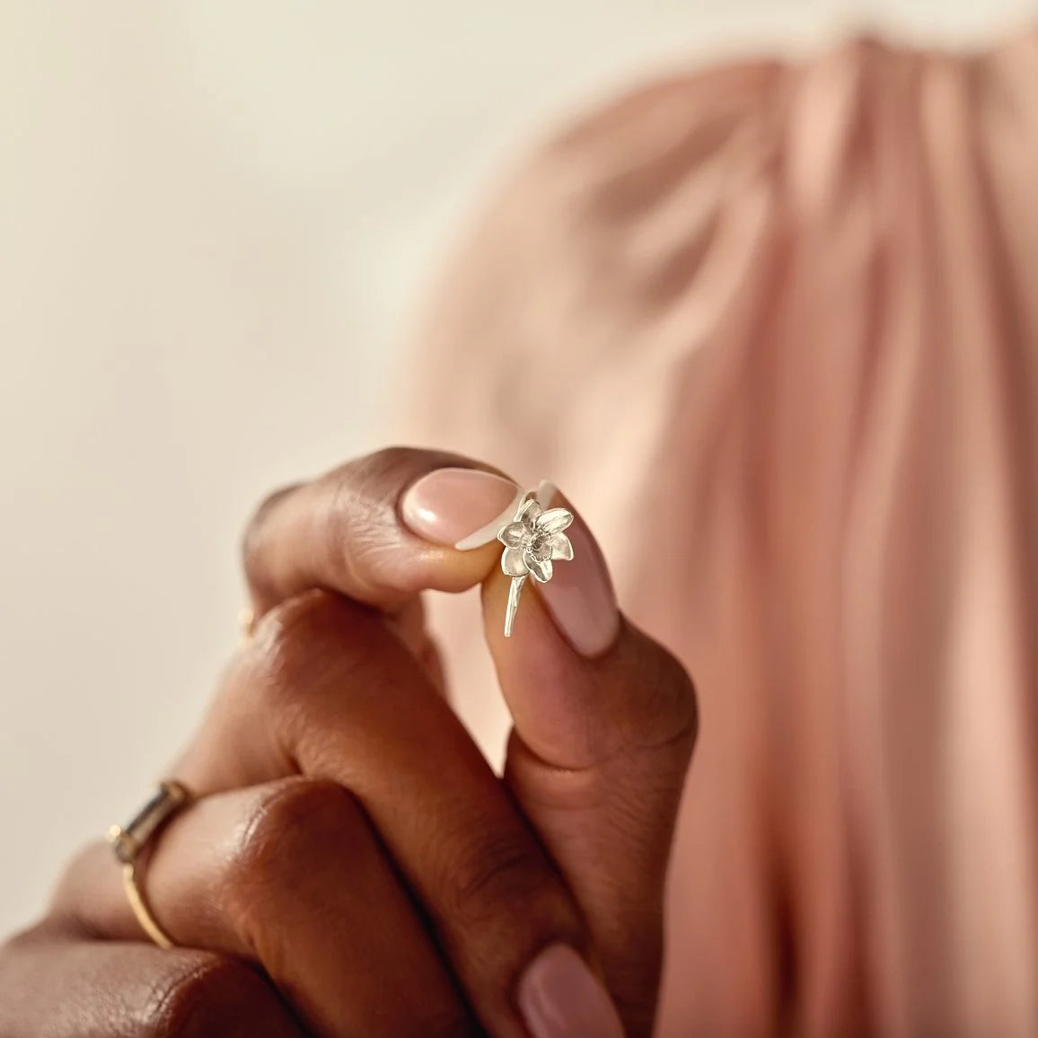 Close-up of a hand wearing a delicate silver ring and a floral charm on a soft pink background.
