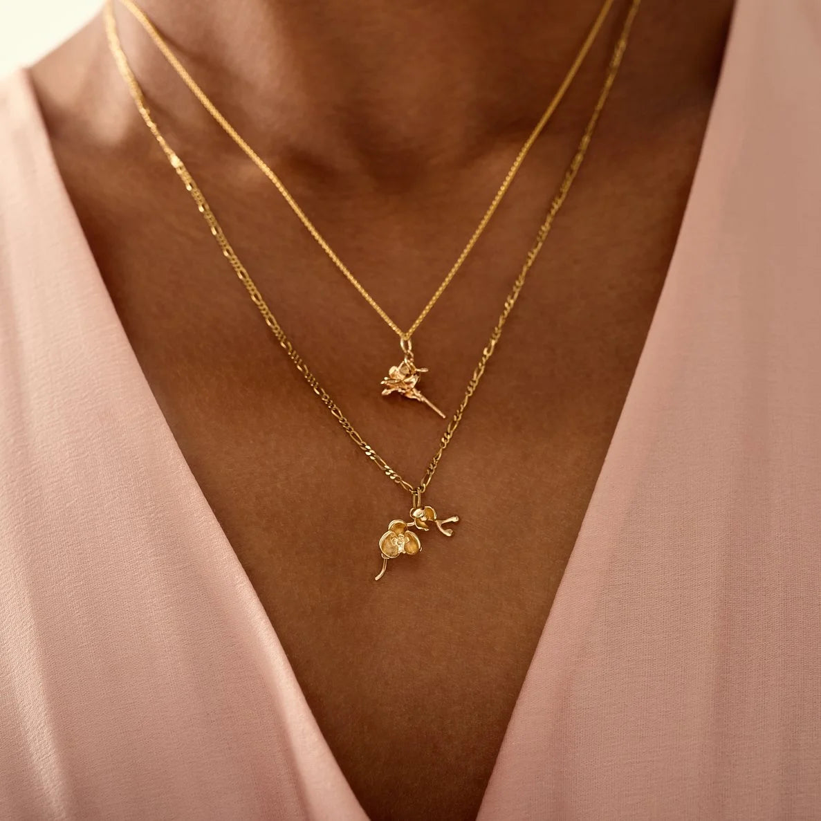 Two gold necklaces with floral pendants on a person wearing a pink top.