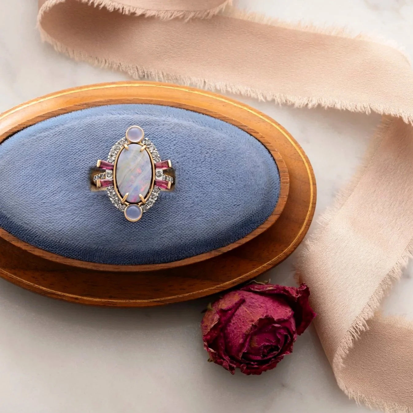 Jewelry box with a opal ring on a blue cushion, next to a dried rose and beige fabric.