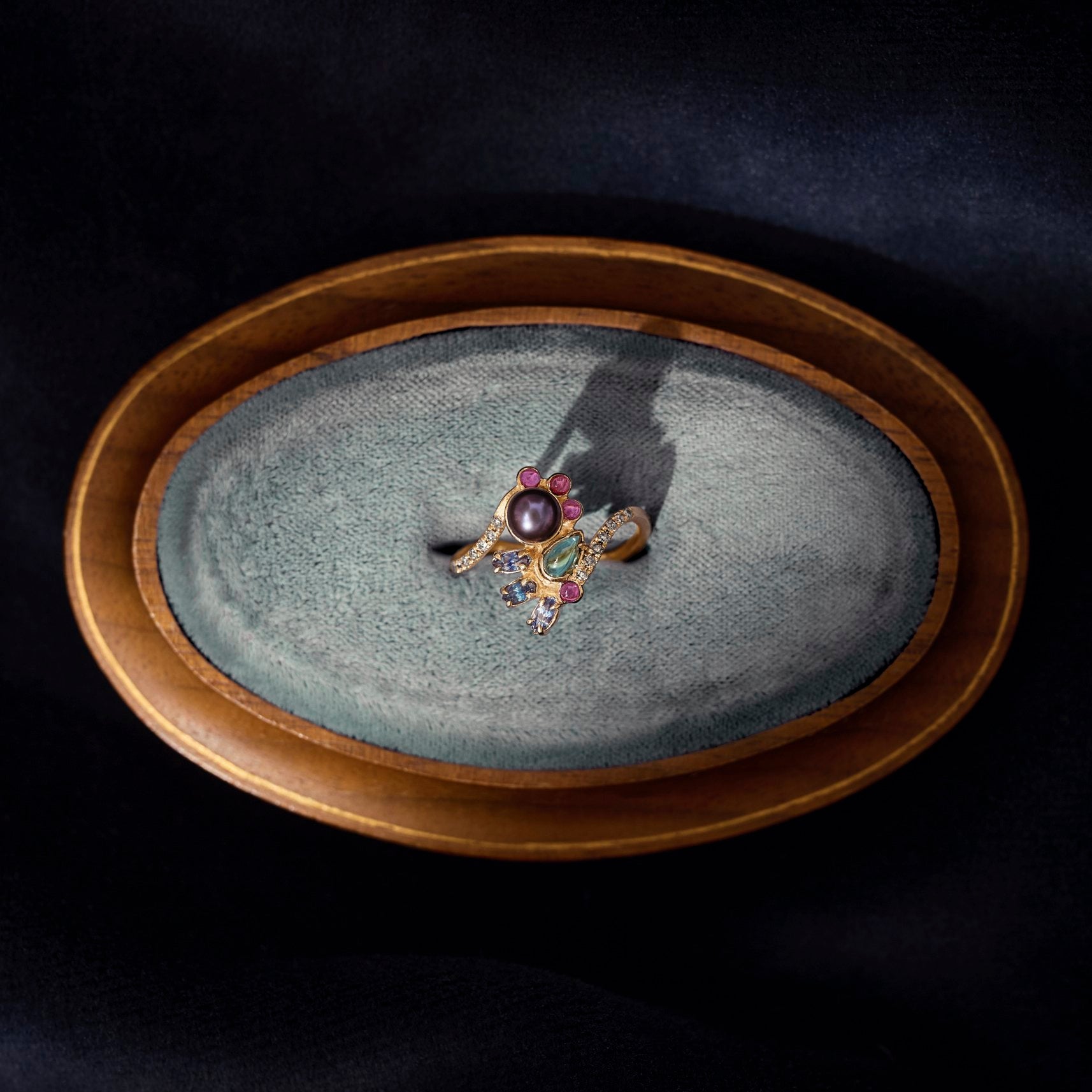 Gold ring with colorful gemstones on a textured stone surface inside an oval wooden frame.