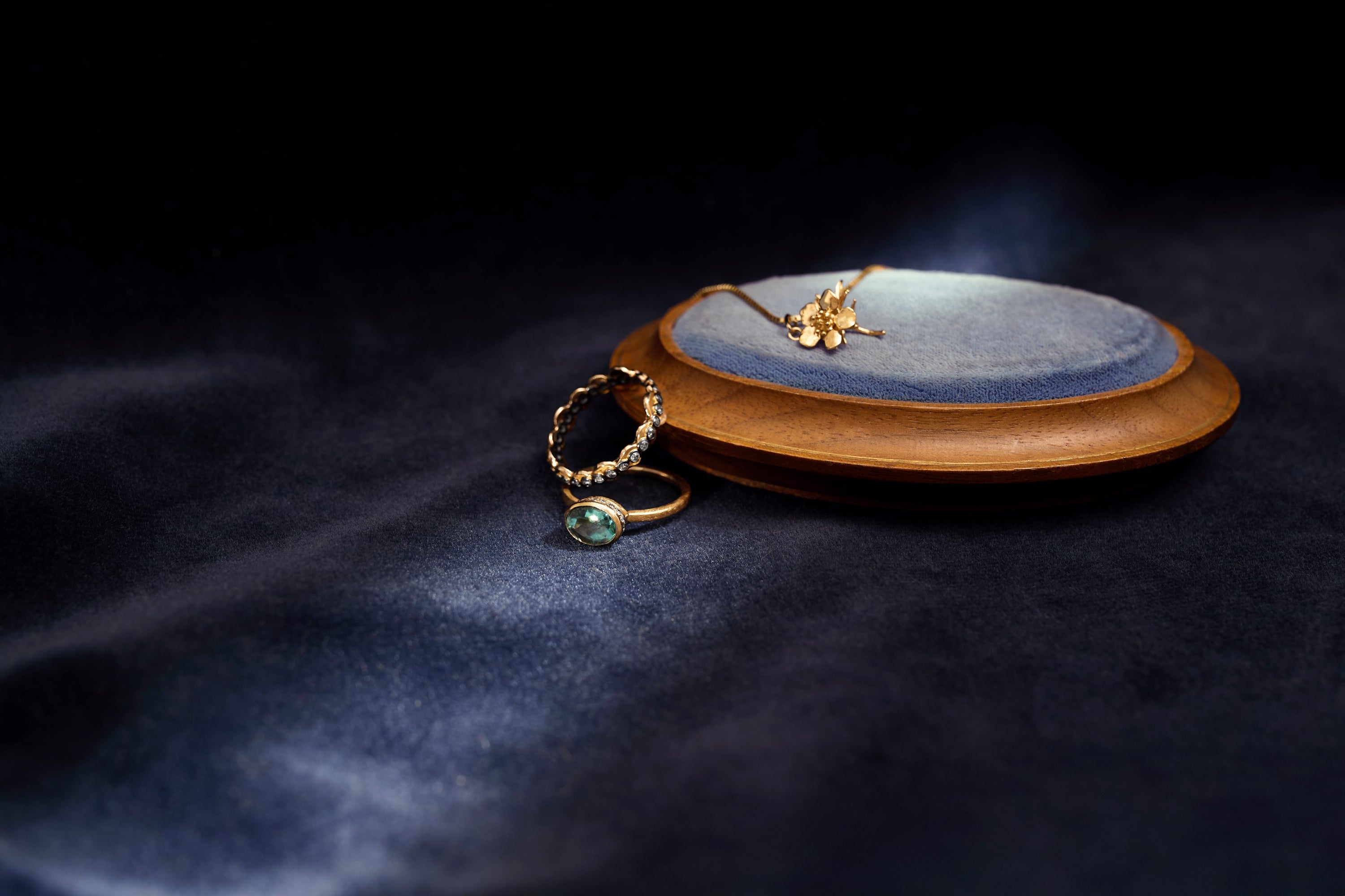 Two gold and gemstone rings on a dark blue fabric background with gold flower charm pendant