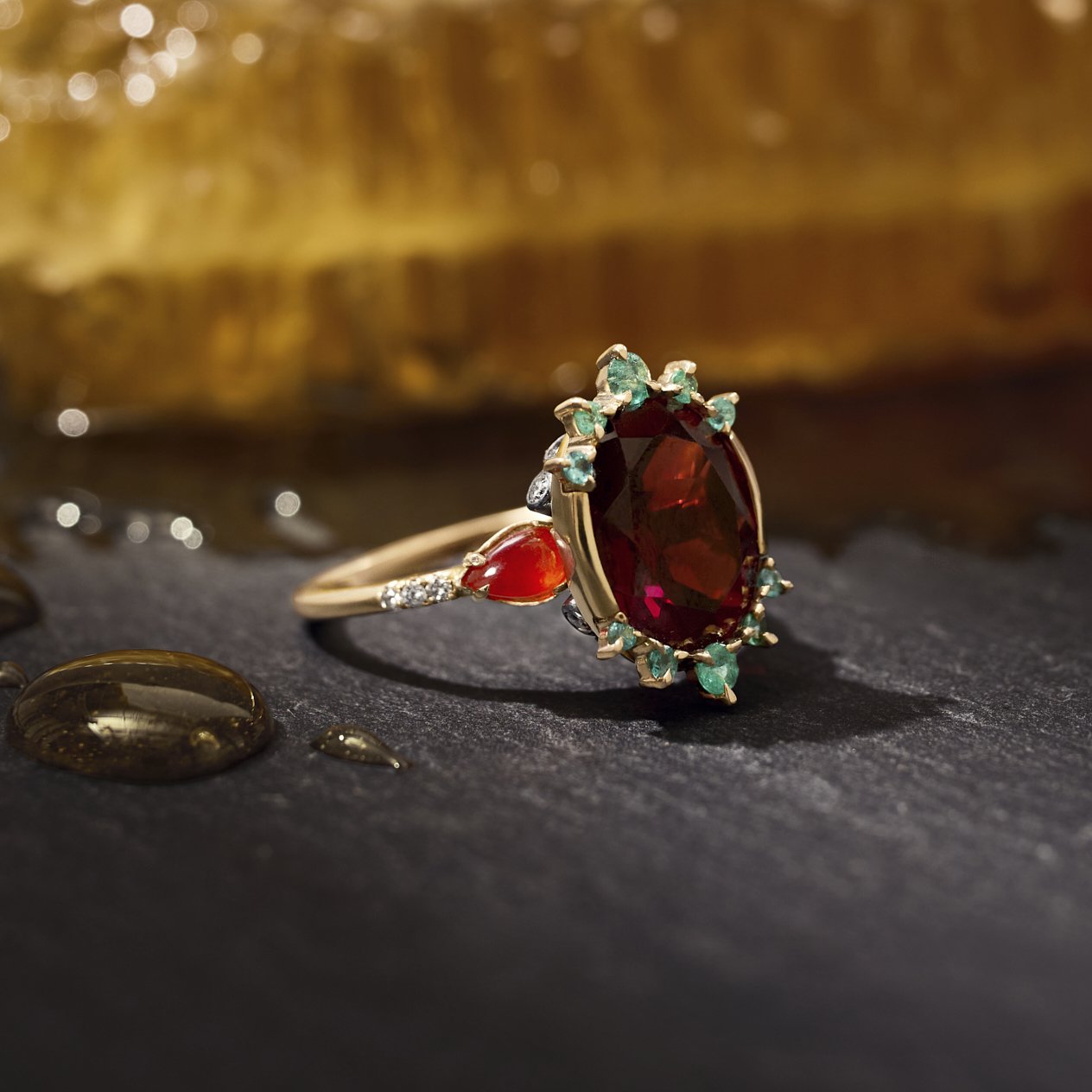 A gold ring with a large oval deep red gemstone, a smaller red teardrop gemstone, and small green accent stones, displayed on a dark textured surface with a blurred golden background.
