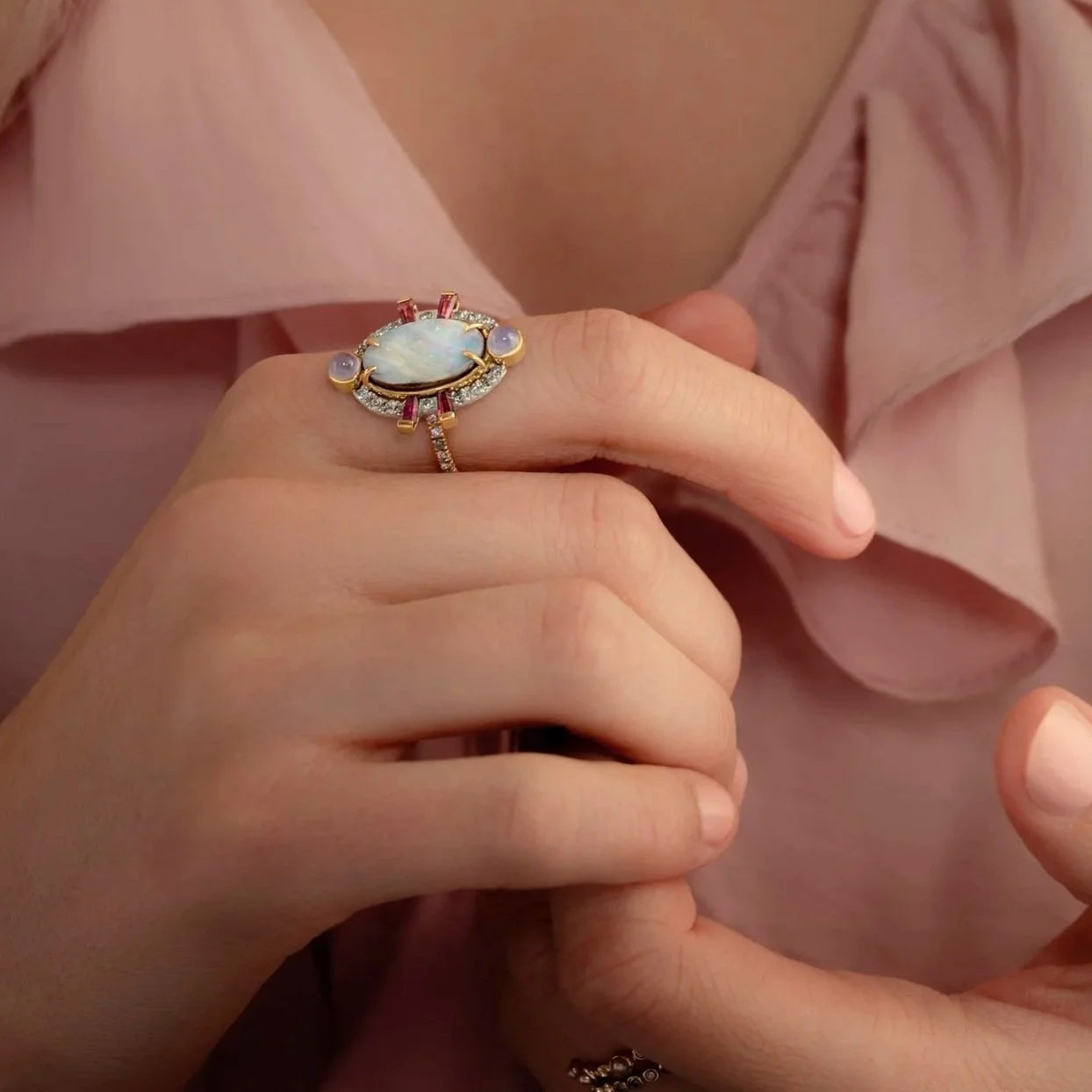 Close-up of a hand wearing an ornate ring with a large gemstone, against a soft pink background.