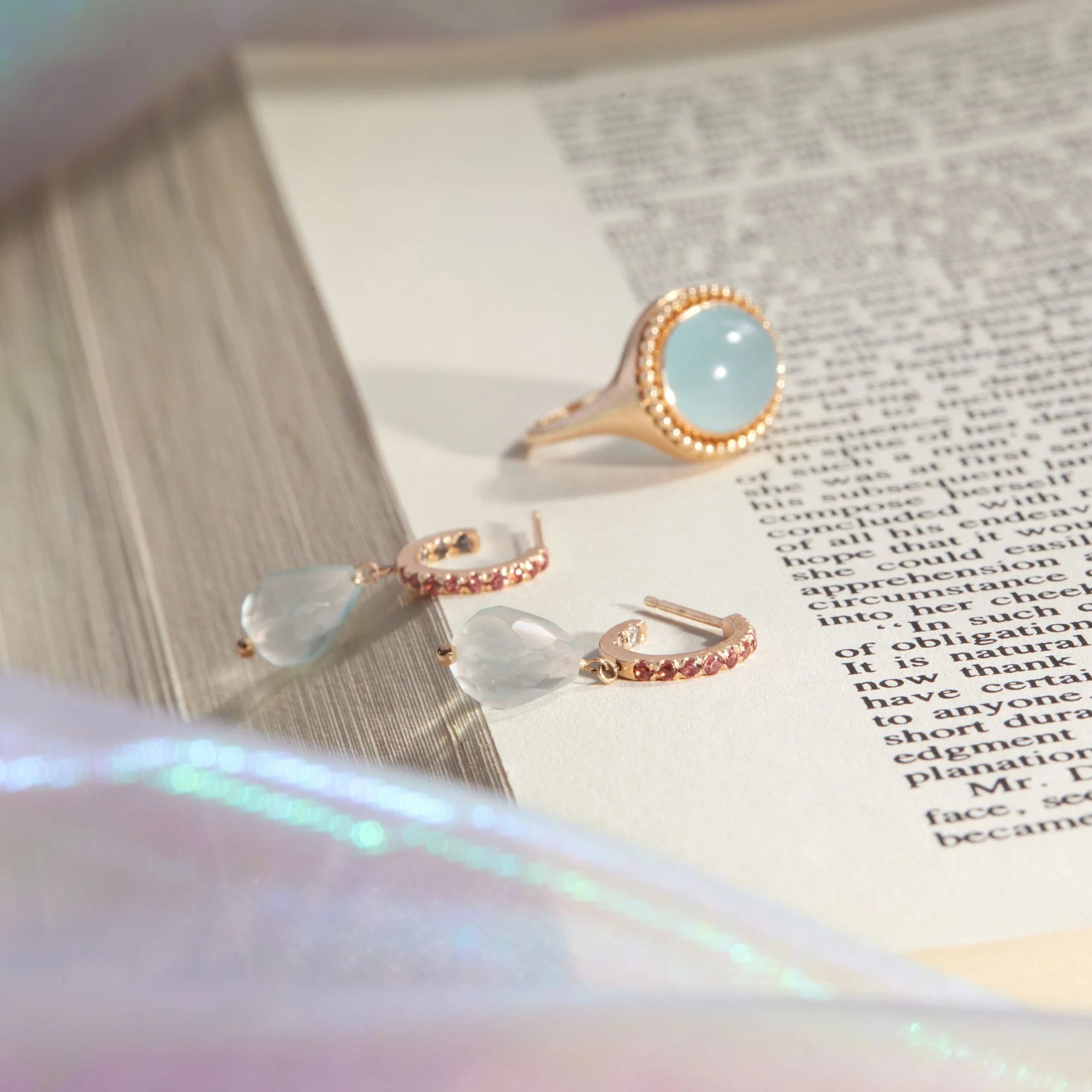 Pair of gemstone encrusted hoop earrings with blue drop and matching gold signet ring with large blue stone on open book.