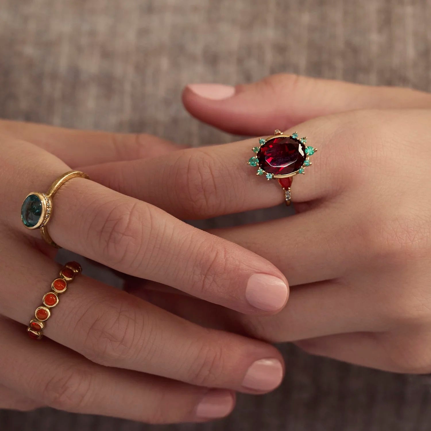 A close-up of a person's hands wearing multiple rings. The most prominent ring features an oval deep red gemstone in a gold setting with small green accents.