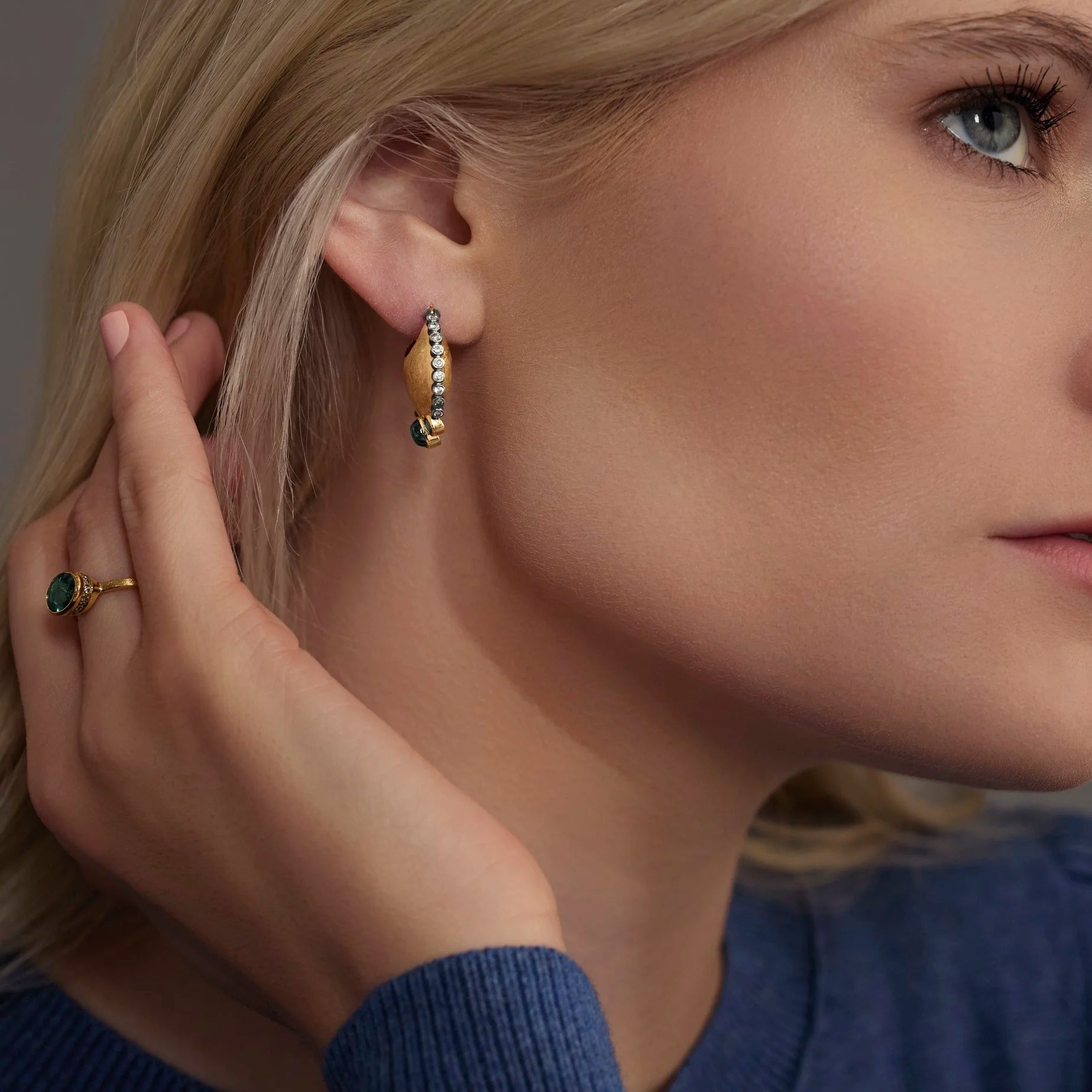 Close-up of a woman wearing gold hoop earrings and a ring with a blue sweater.