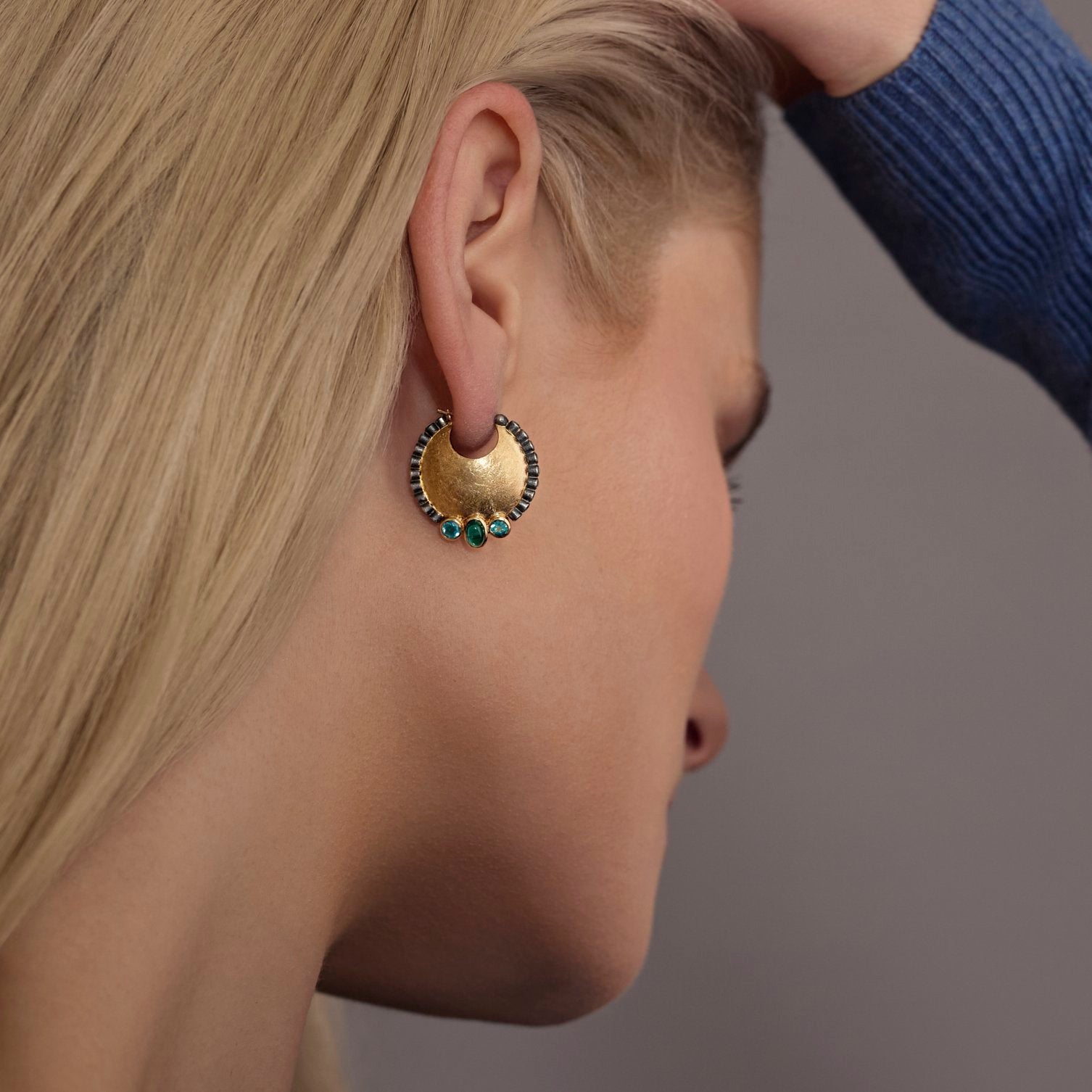 Close-up of a person wearing a gold earring with teal stones on a neutral background