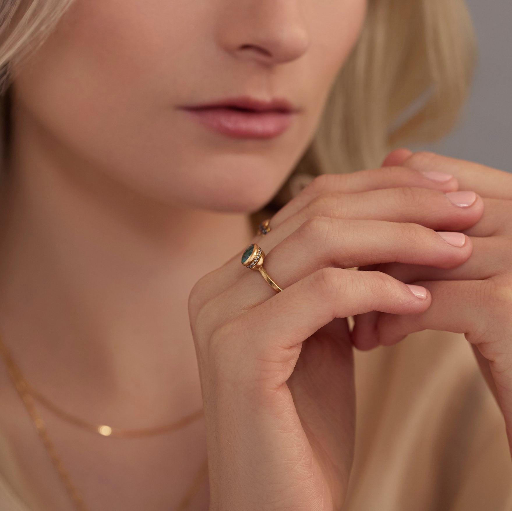 Close-up of a woman's hand wearing a gold ring with a teal gemstone, against a neutral background.