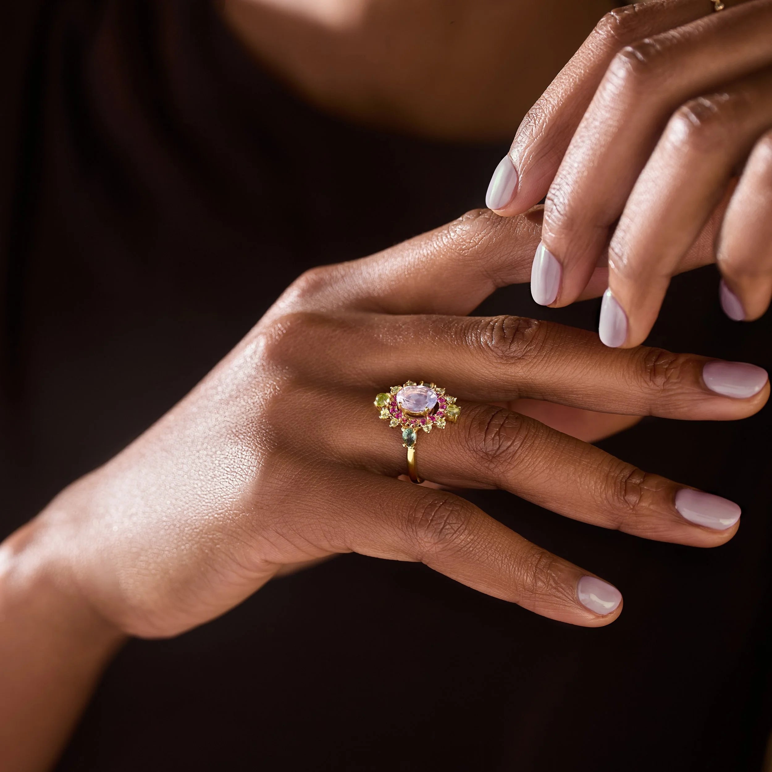 A close-up lifestyle shot of a gold ring with a central lavender-purple oval stone and surrounding pink and green gemstones, worn on a person's finger.