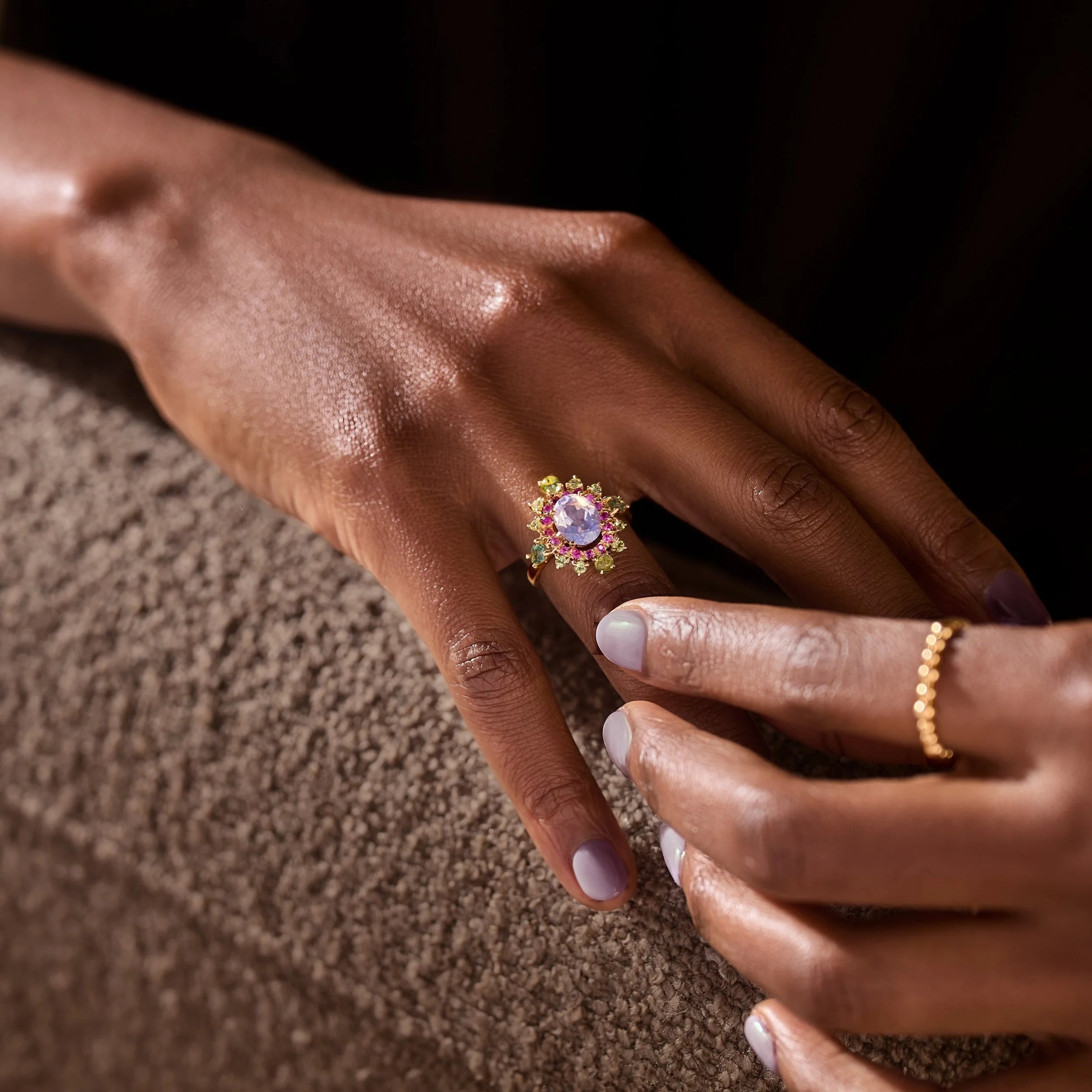 A close-up lifestyle shot of a hand wearing a gold ring with a central lavender-purple gemstone, while another hand gently touches it. The background is a dark, textured brown surface.