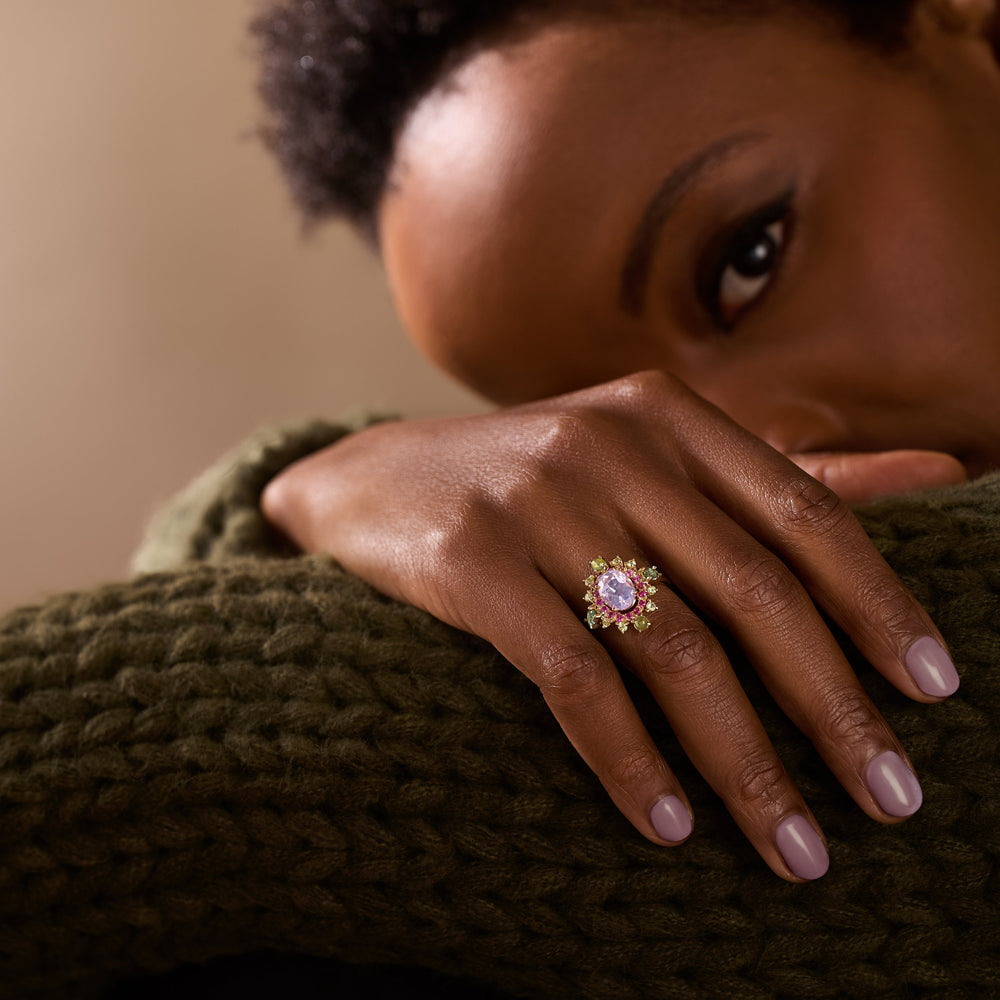 A close-up lifestyle shot of a model wearing a gold ring with a lavender-purple oval center stone and small yellow accent stones, resting her hand on a chunky olive green knit sweater.