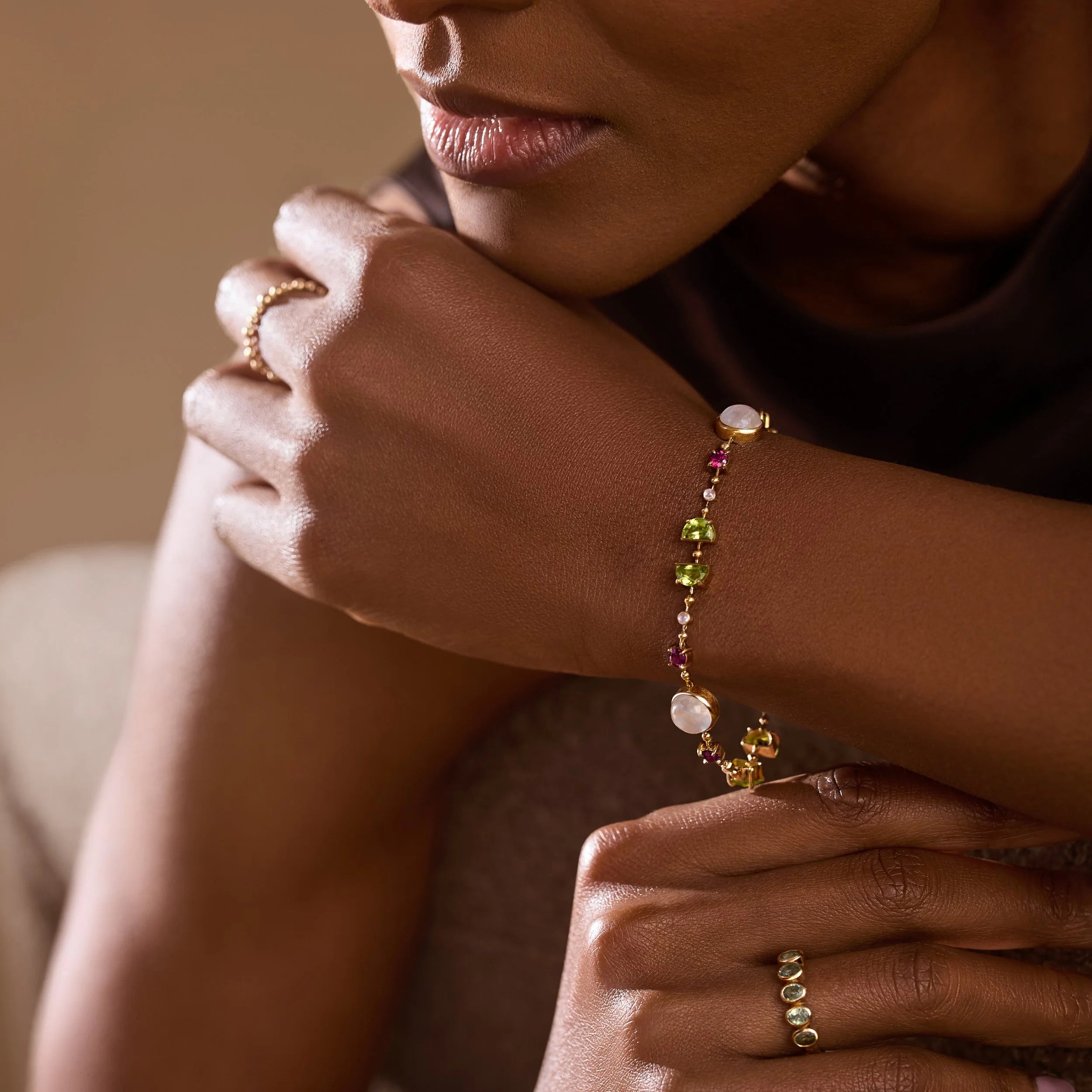 Close-up of a person wearing a gemstone bracelet and rings with a warm, brownish background.