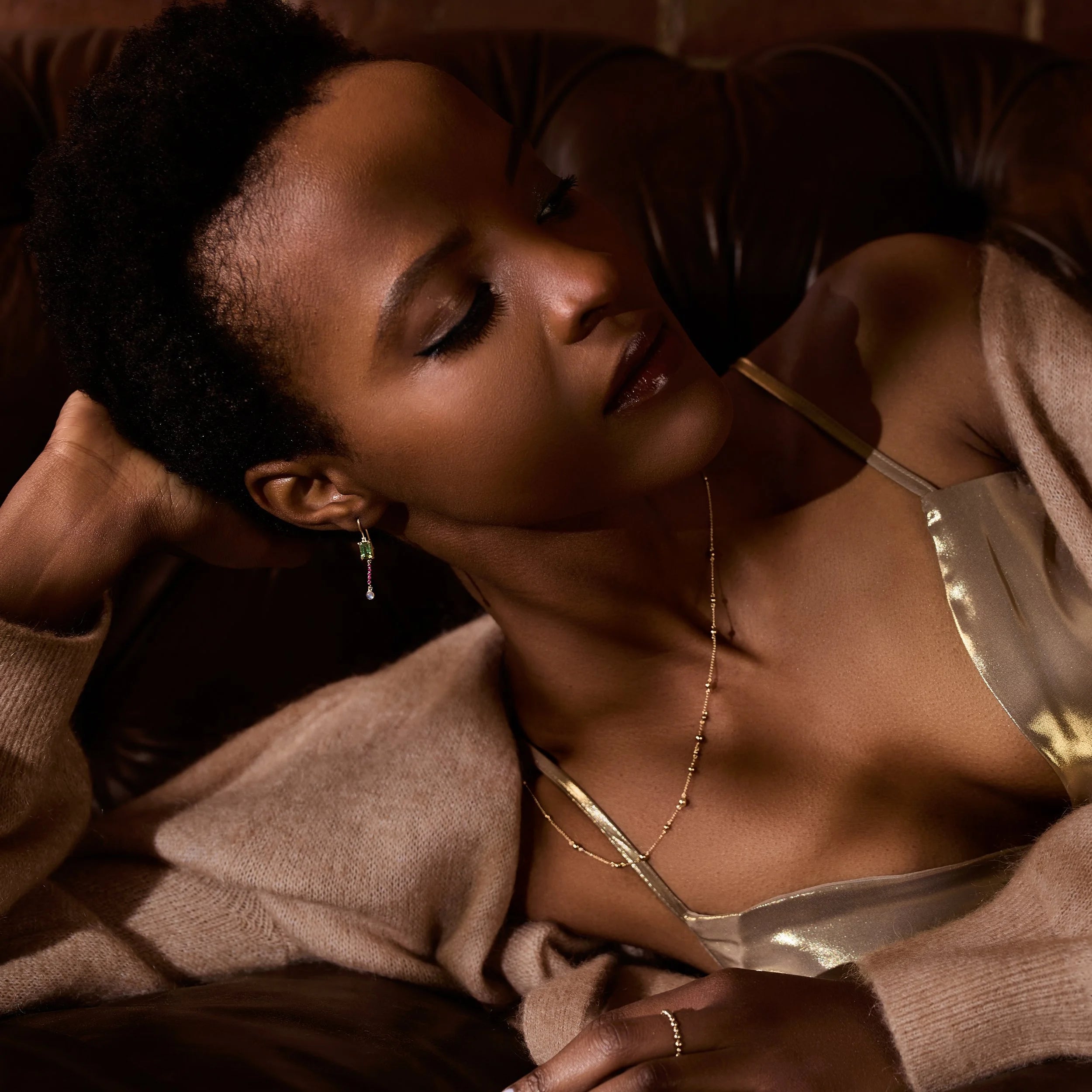 Woman wearing a gold jewelry and dress and beige sweater in a warm-toned setting