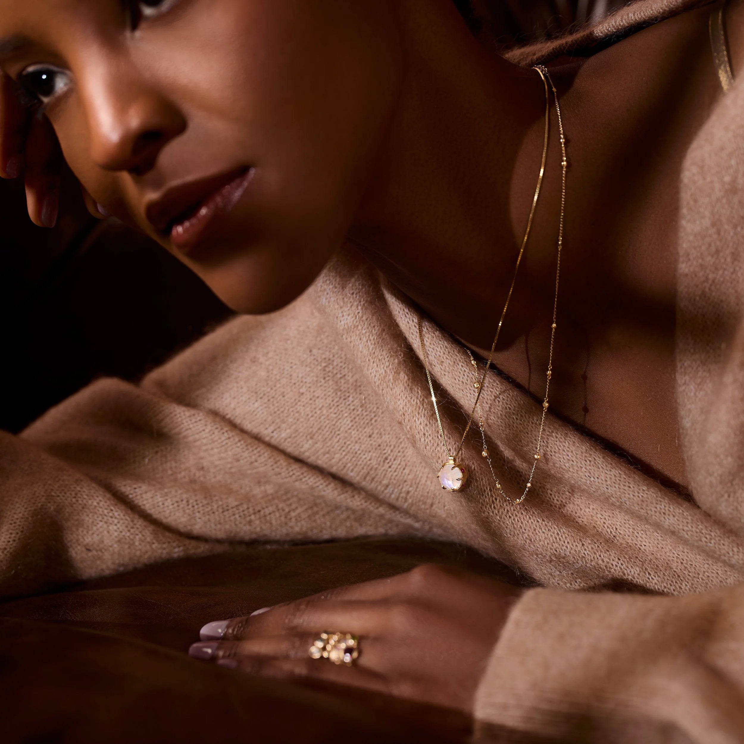 Woman wearing gold jewelry including a necklace and ring, with a warm-toned background.