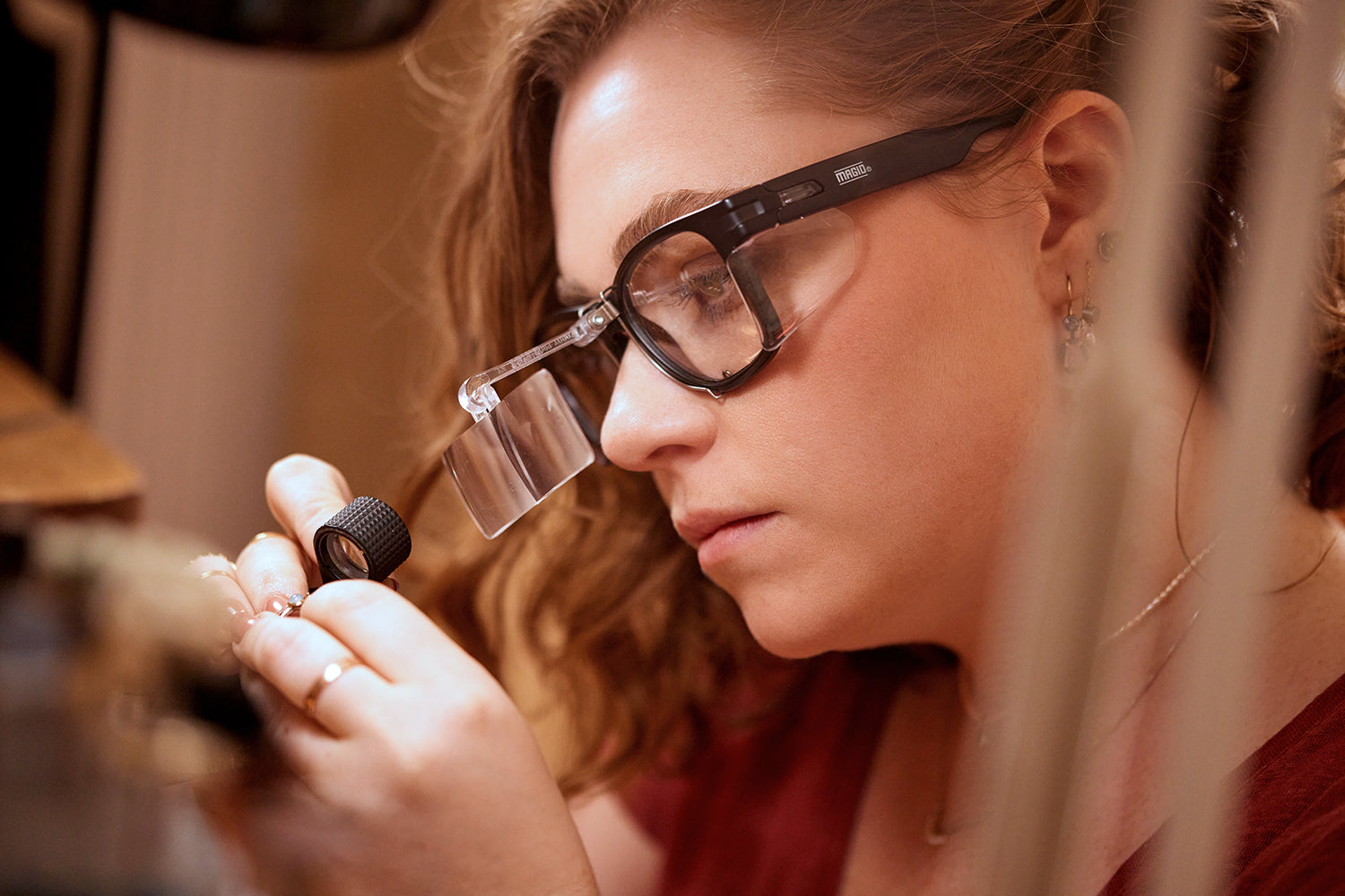 Person wearing glasses examining a small object with a magnifying glass.