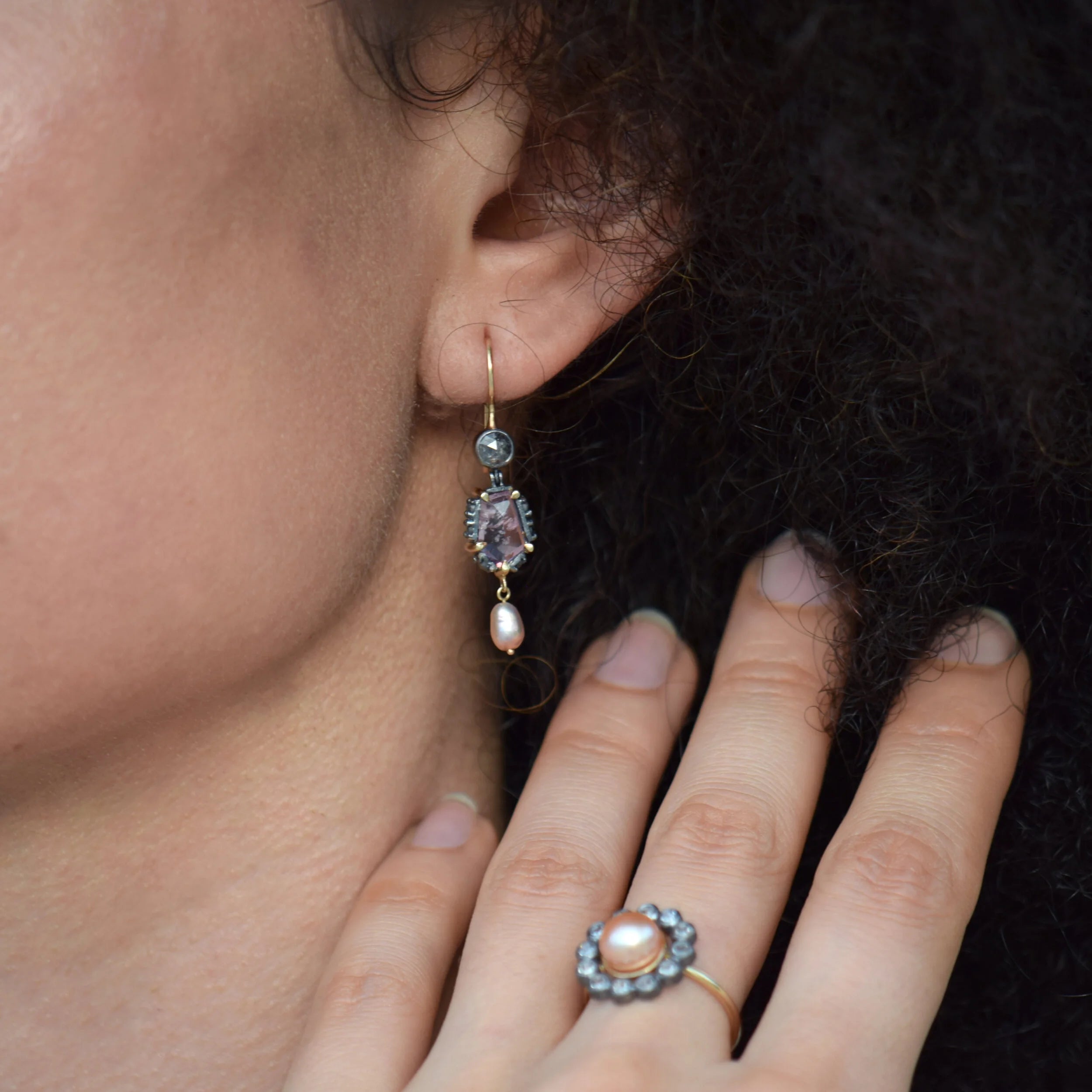 Close-up of an ear wearing a dangling earring with a hand holding a ring featuring similar jewelry design.