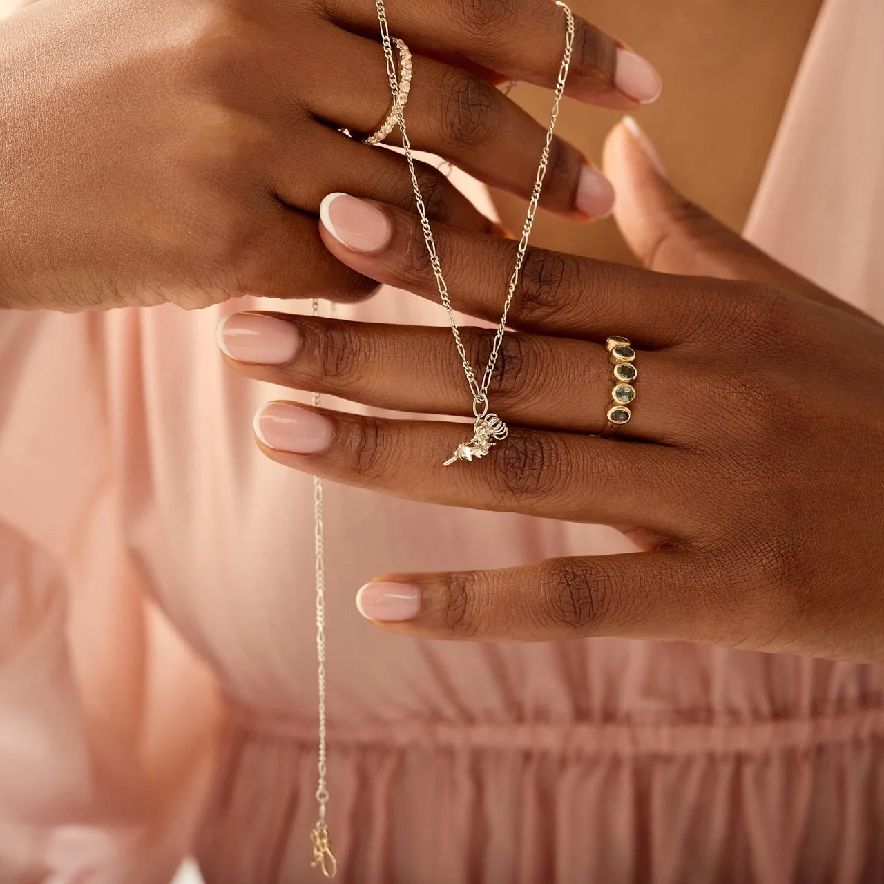 Close-up of hands wearing gold rings with a necklace on a soft pink background