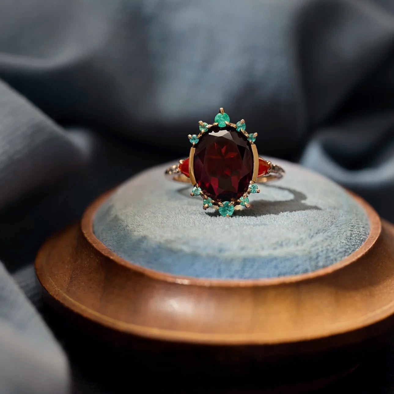 An antique-style gold ring with a large oval dark red garnet center stone and small green gemstones, displayed on a textured blue-gray ceramic dish over a wooden surface.