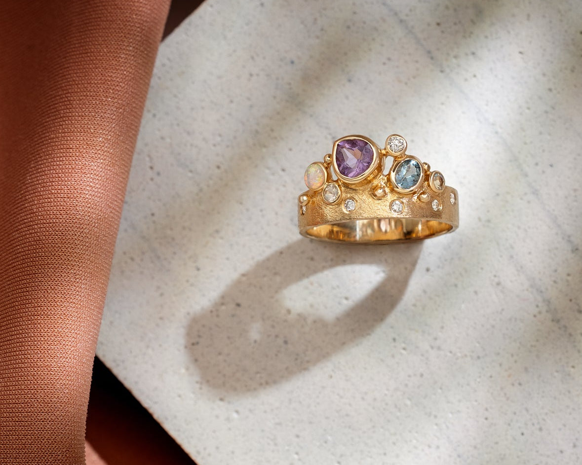 Gold ring with multiple gemstones in purple and blue with diamonds on tile and brown fabric