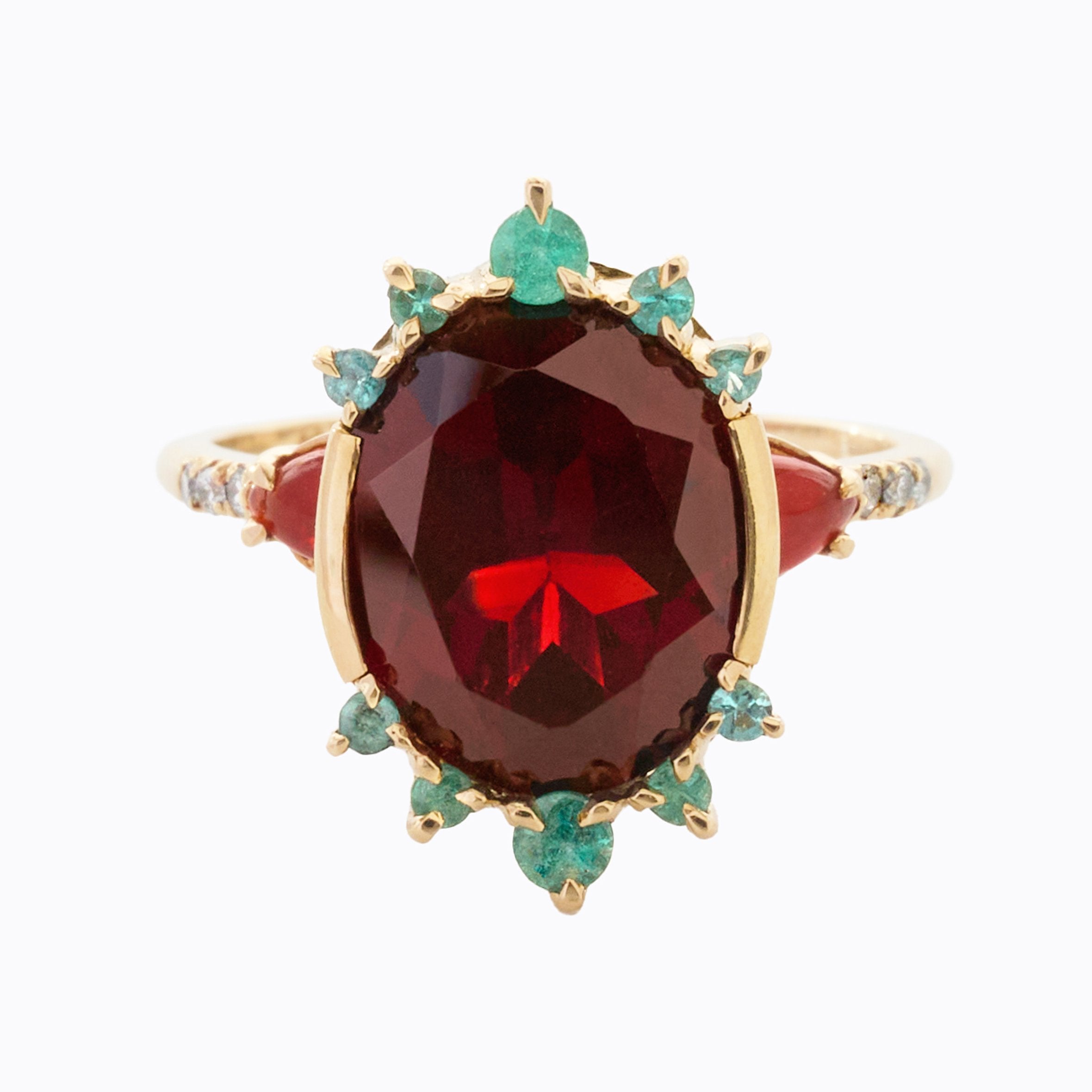 A gold ring featuring a large oval deep red gemstone, accented by small teal stones and tiny diamonds on the band, set against a plain white background.