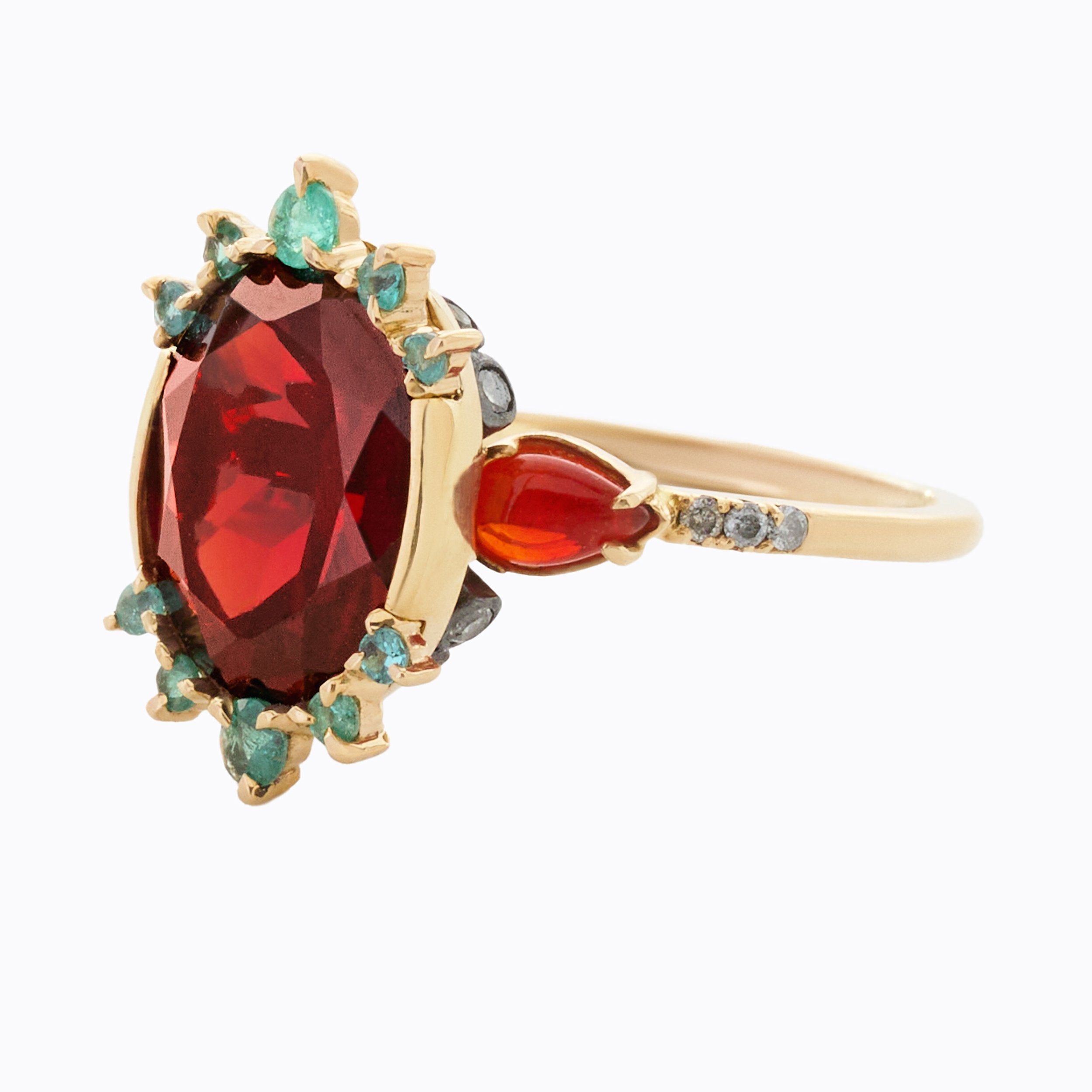 A gold ring featuring a large oval red gemstone surrounded by small turquoise and diamond stones, with a smaller orange gemstone on the band and tiny diamond accent stones.