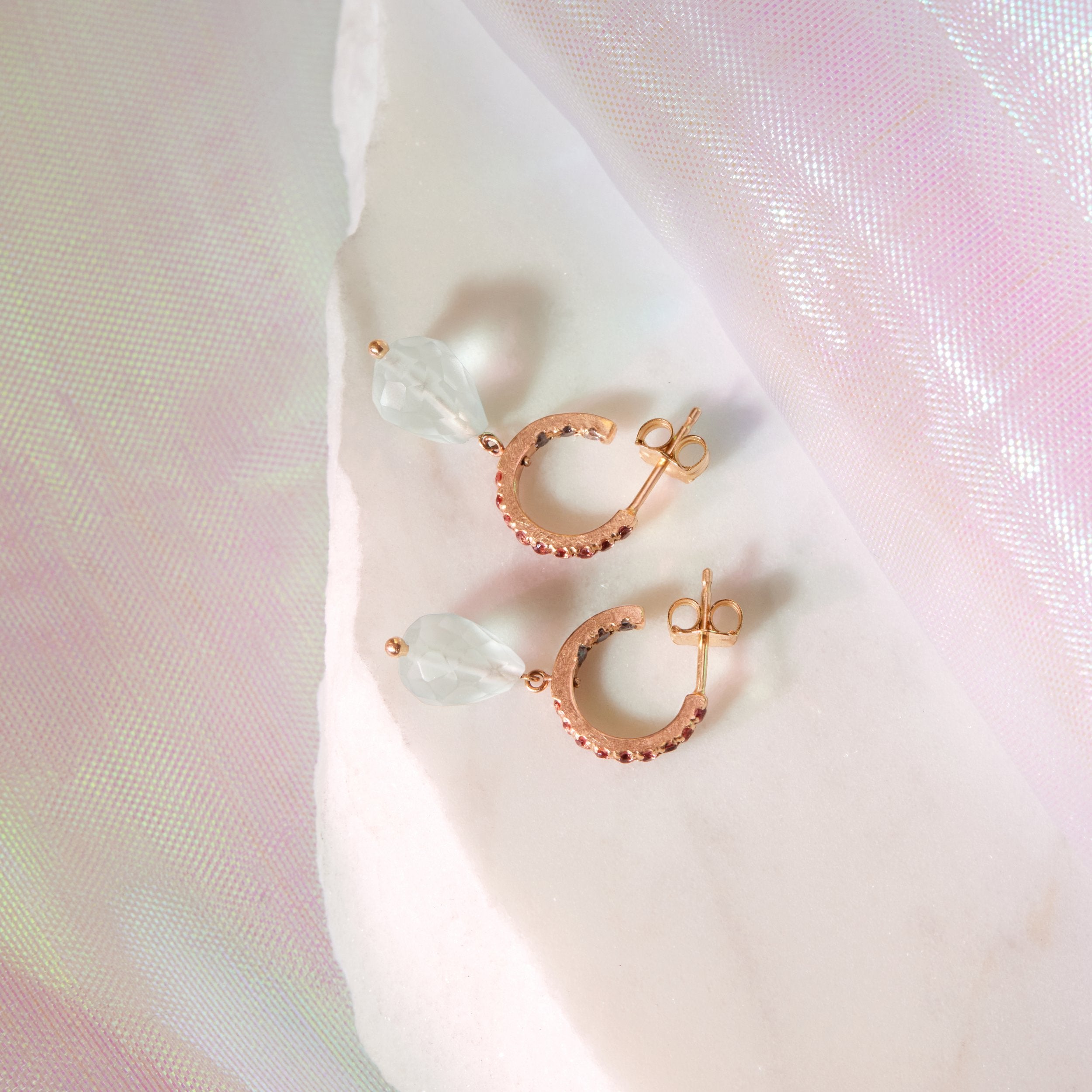 Gold huggie hoops with pink and gray stones and blue gemstone drop on white fabric.