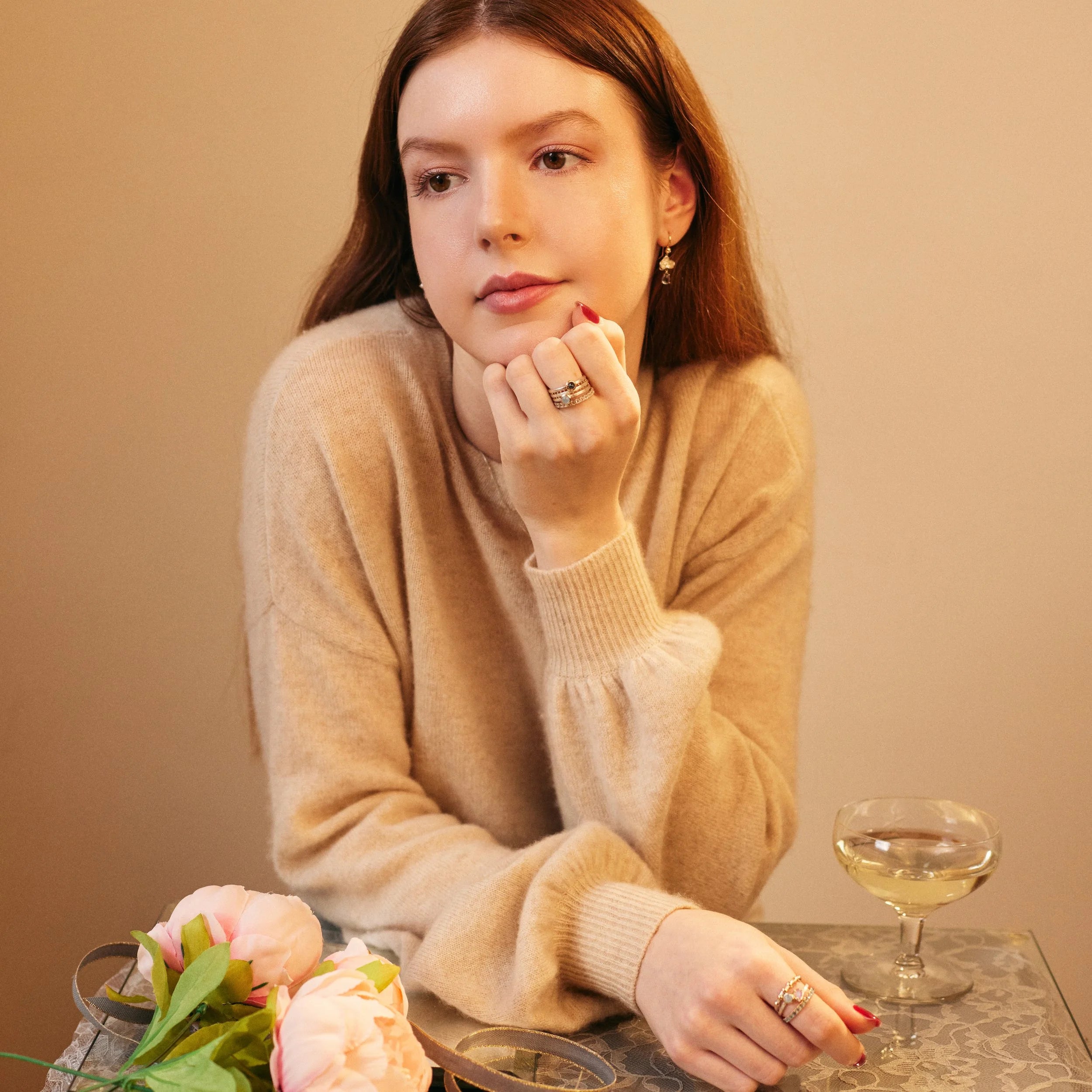 Woman in a beige sweater sitting at a table with flowers and a glass of wine, against a beige background