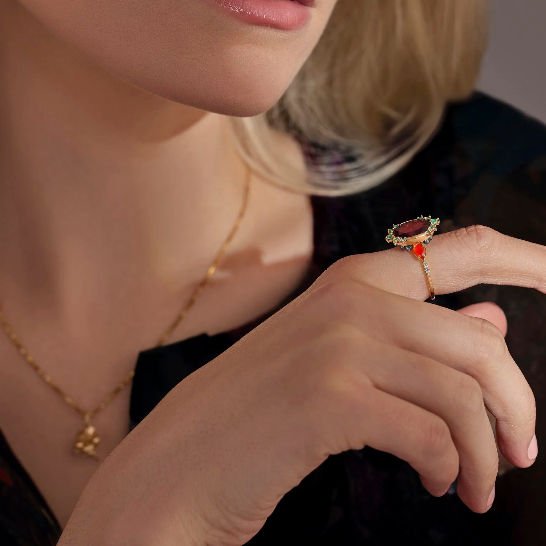 A close-up lifestyle shot of a model wearing a gold ring with a red oval stone and small green and white accents, along with a delicate gold necklace.