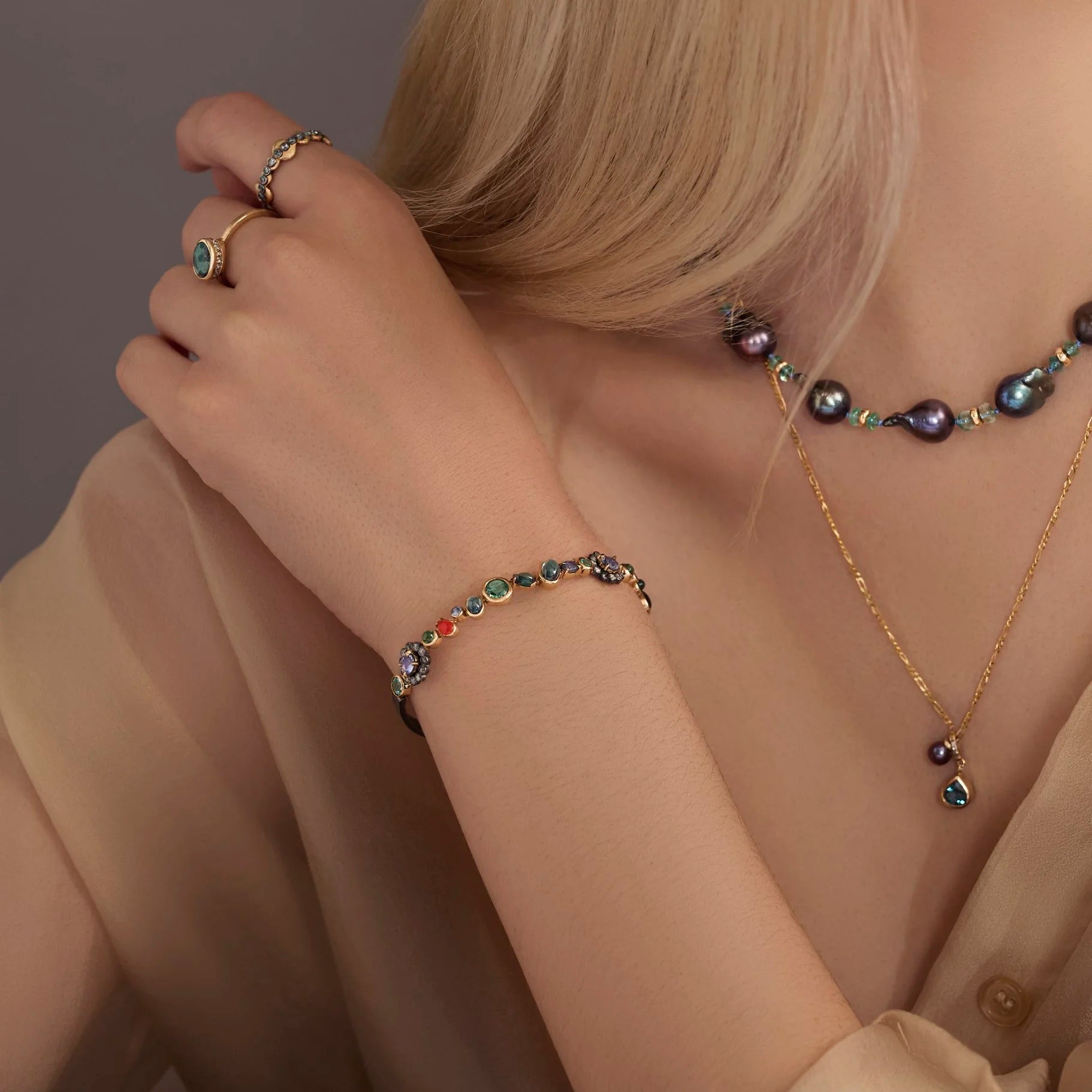 Person wearing a gold bracelet and necklace with colorful gemstones against a neutral background