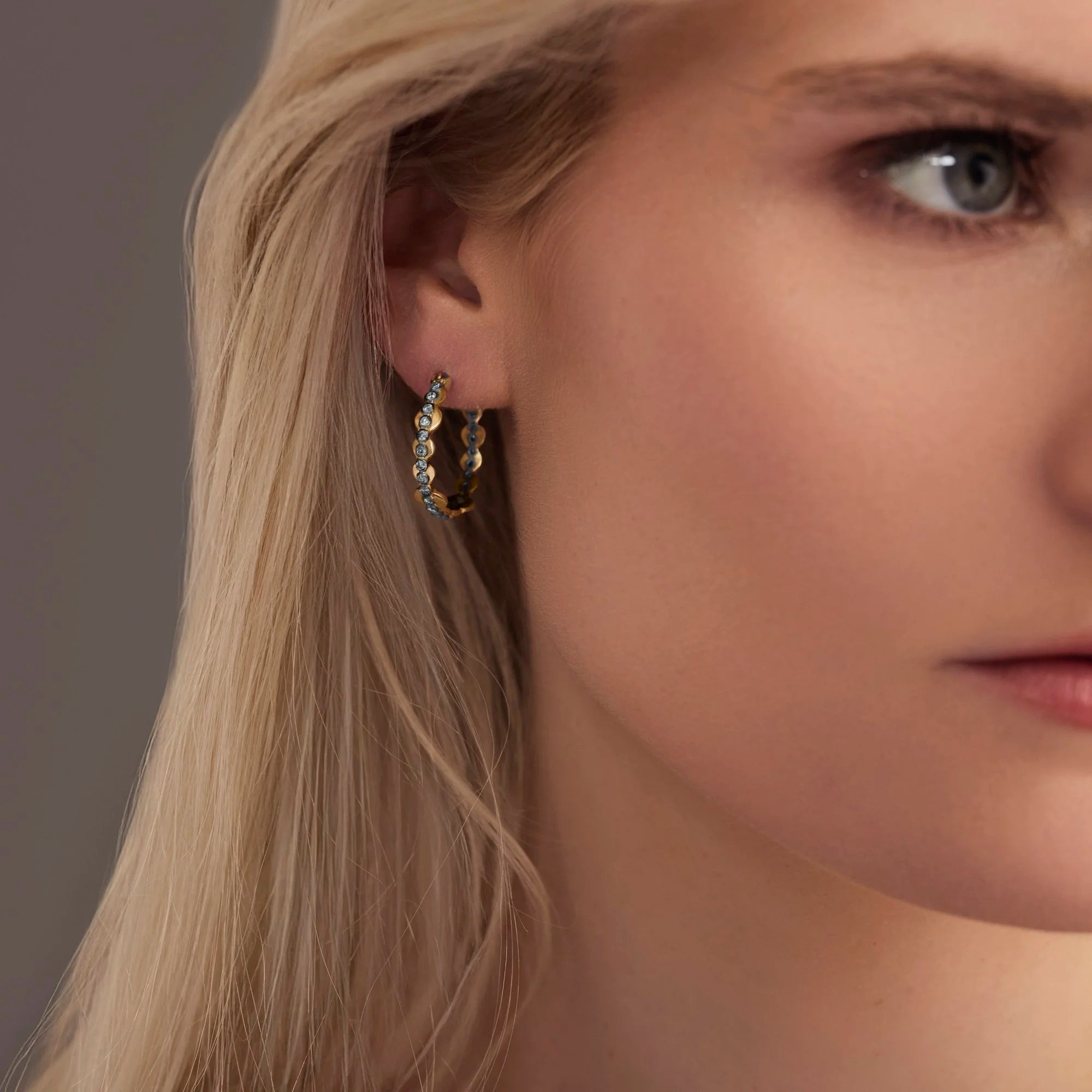 Close-up of a woman wearing gold hoop earrings with embedded stones against a neutral background