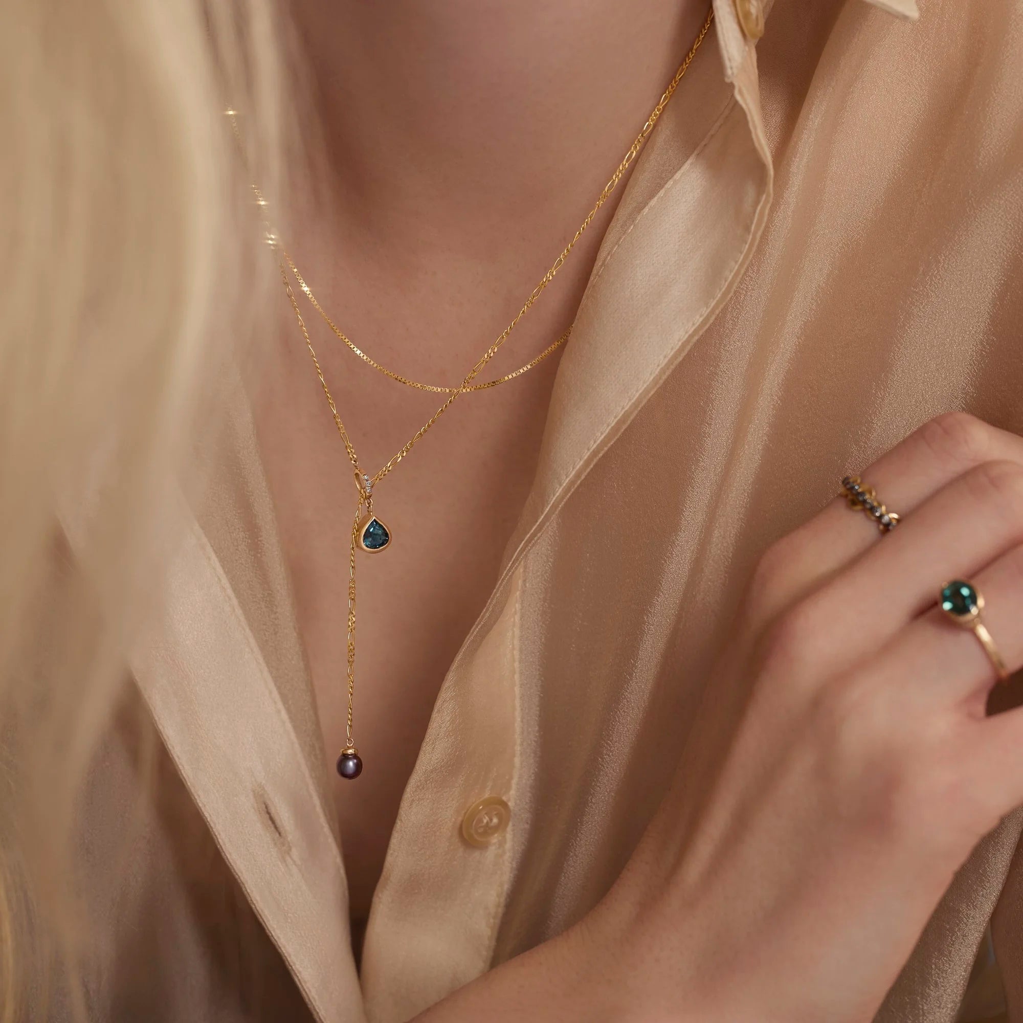 Close-up of a person wearing a gold necklace with gemstones and a matching ring on a beige shirt.