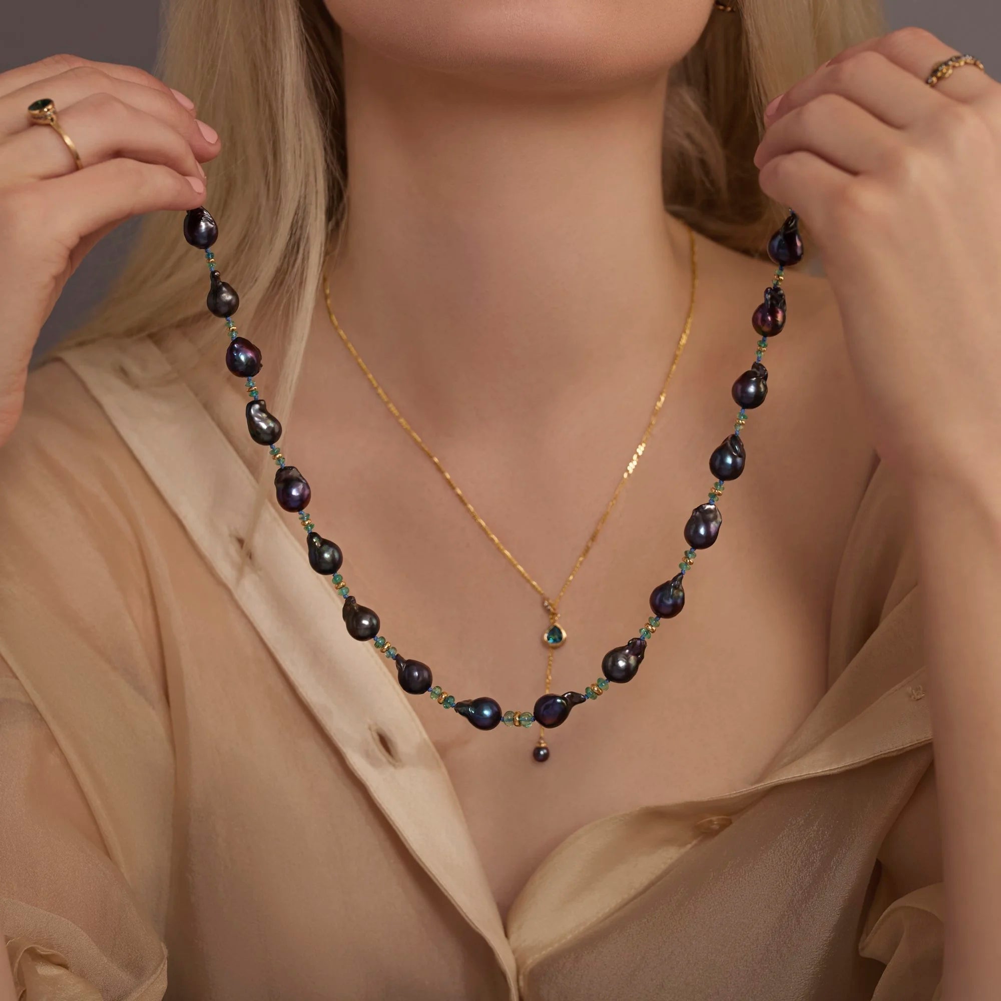 Woman wearing a gold necklace with a black pearl necklace over a beige blouse.