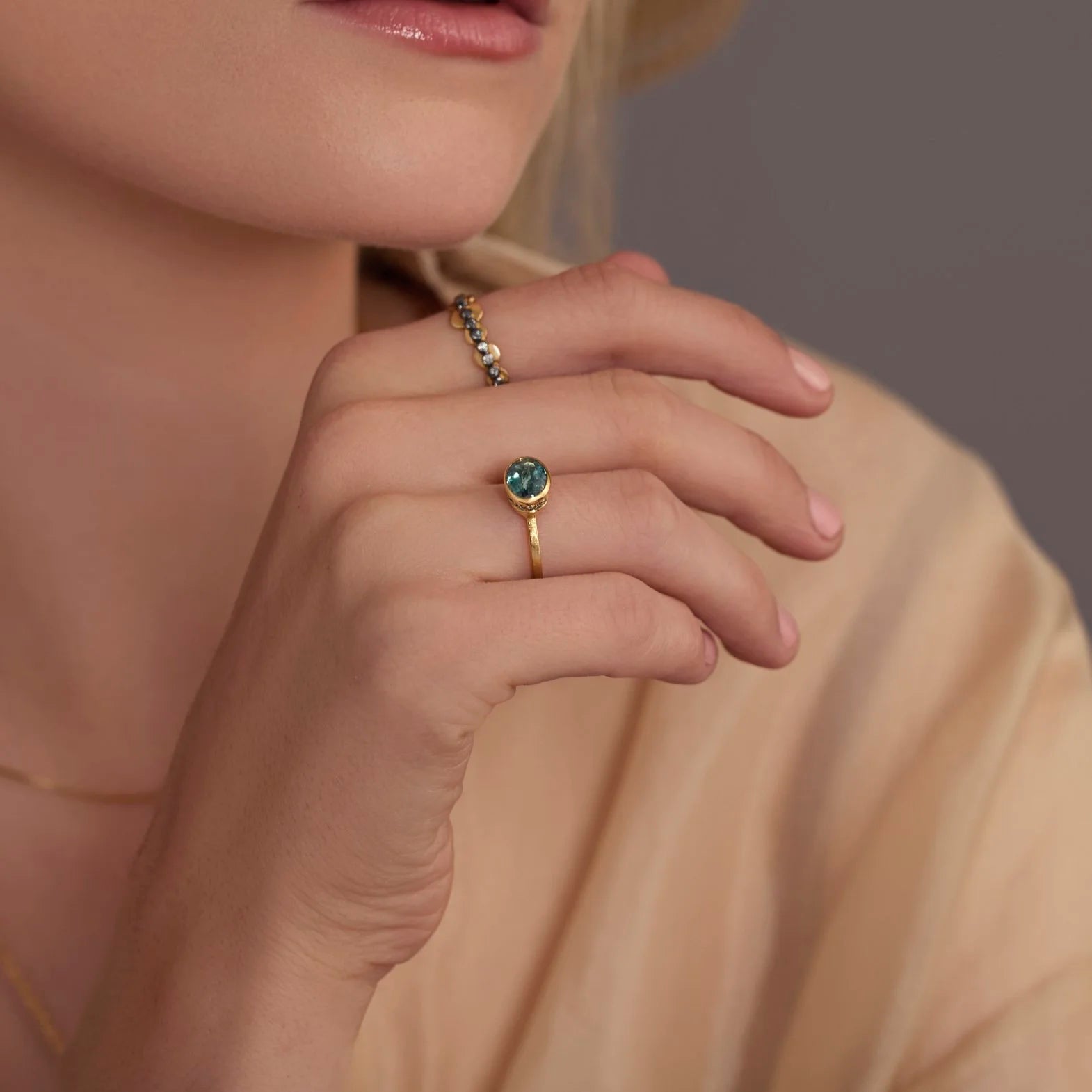 Close-up of a hand wearing a gold ring with a teal gemstone on a neutral background