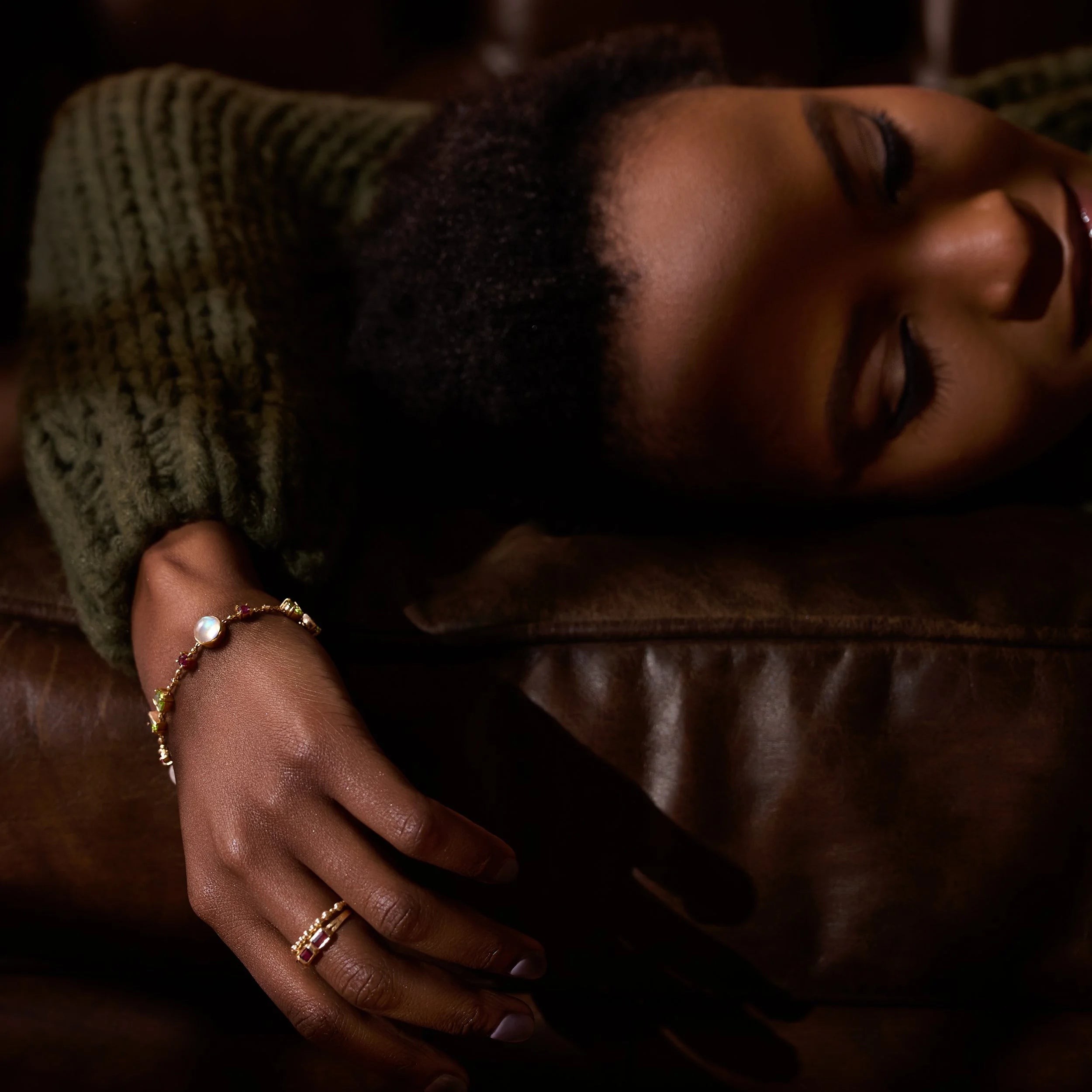 Woman resting her head on a brown surface with gold jewelry on her hand.