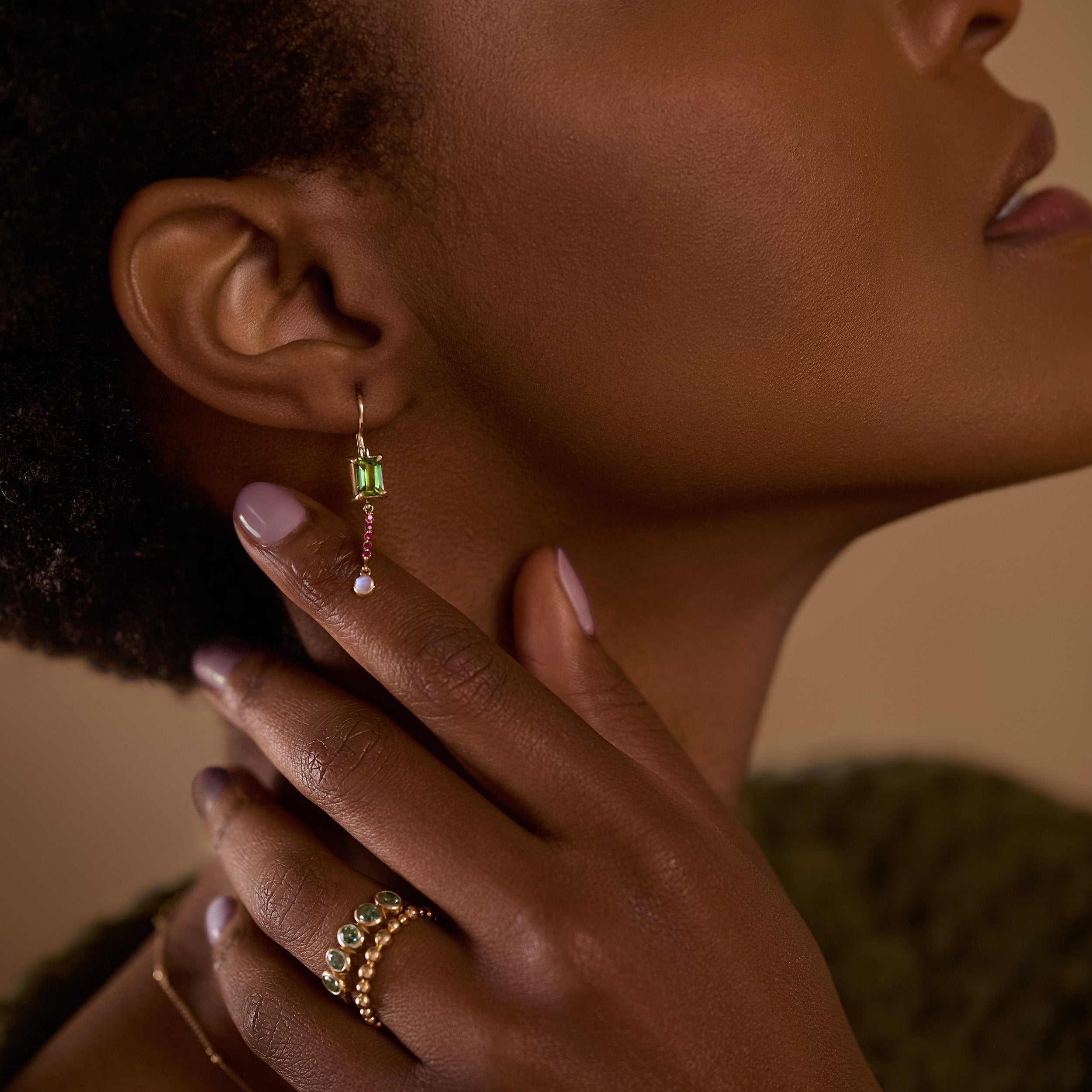 Close-up of a woman wearing gold earrings and rings with green gemstones.