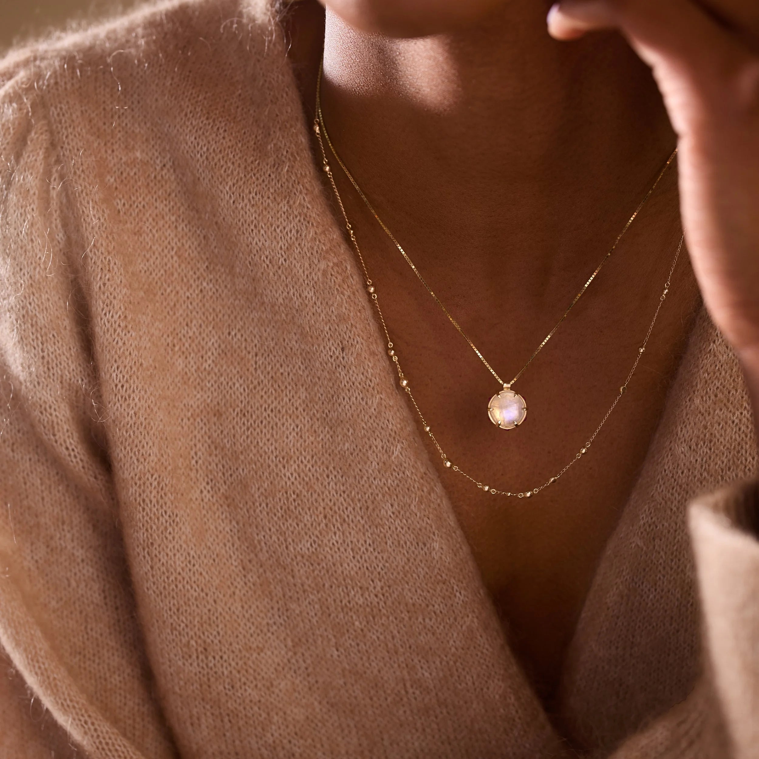 Close-up of a person wearing a gold necklace with a pendant on a beige sweater.