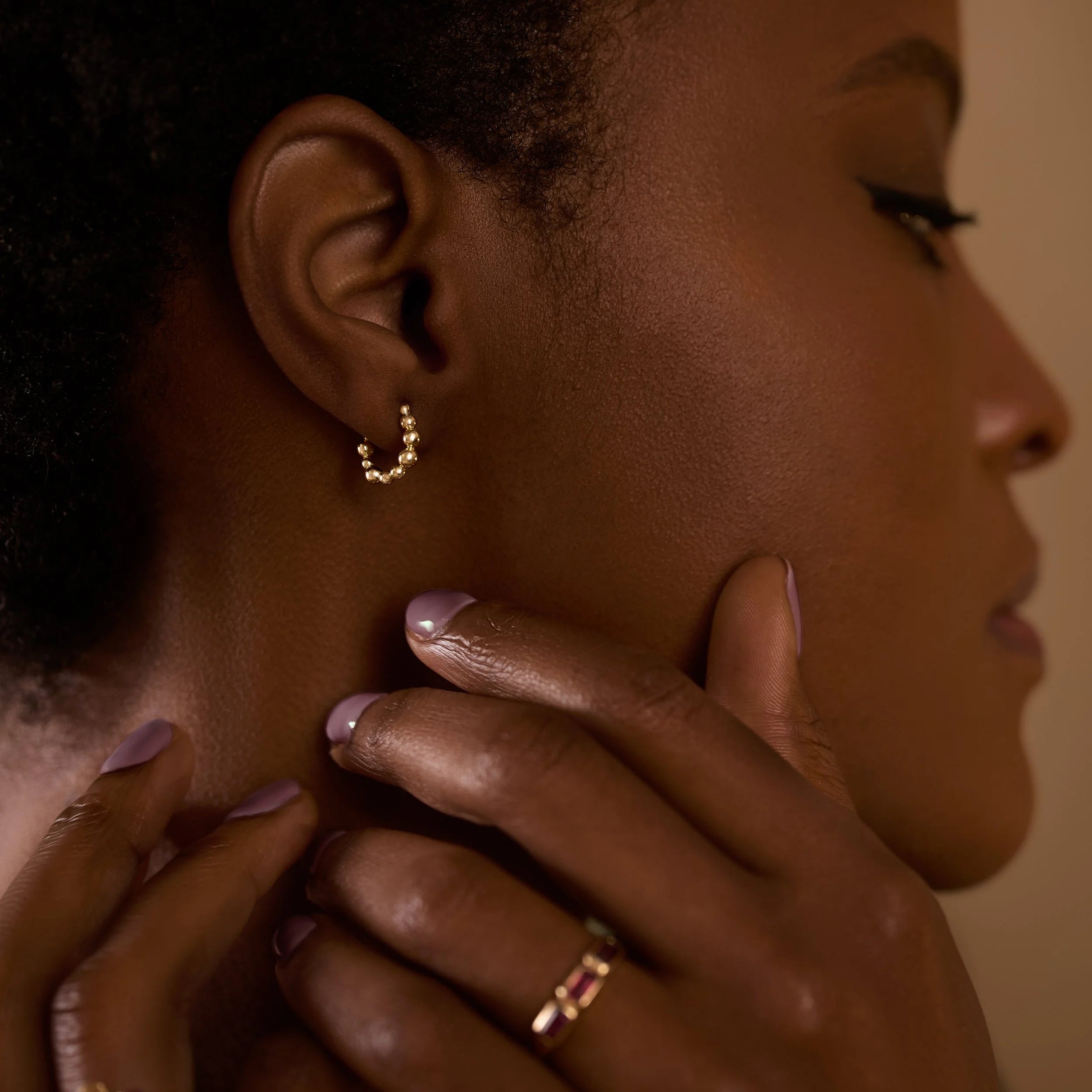 Close-up of a woman wearing gold hoop earrings and a ring, with one hand touching her neck.