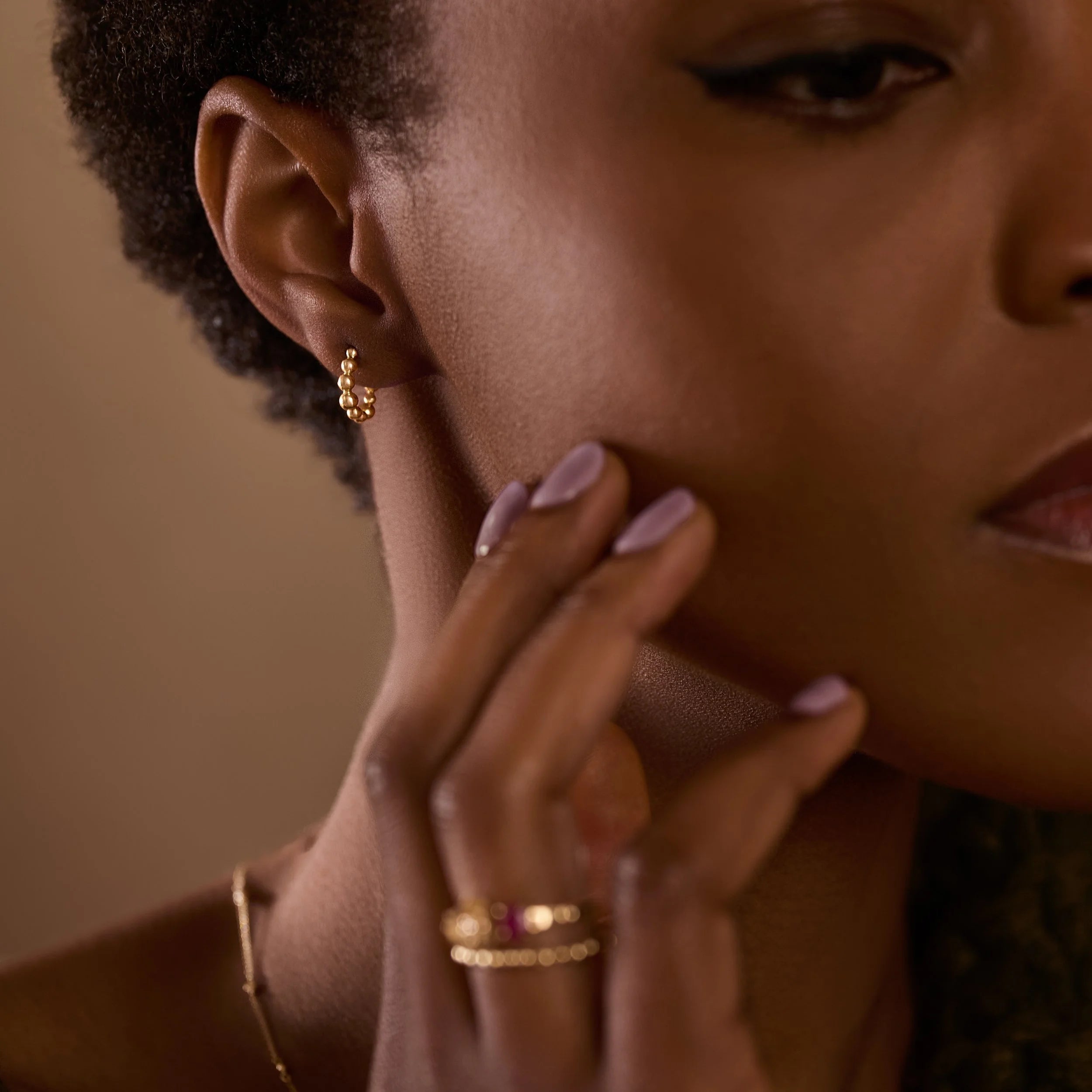 Close-up of a woman's face with gold earrings and rings, wearing a neutral background.