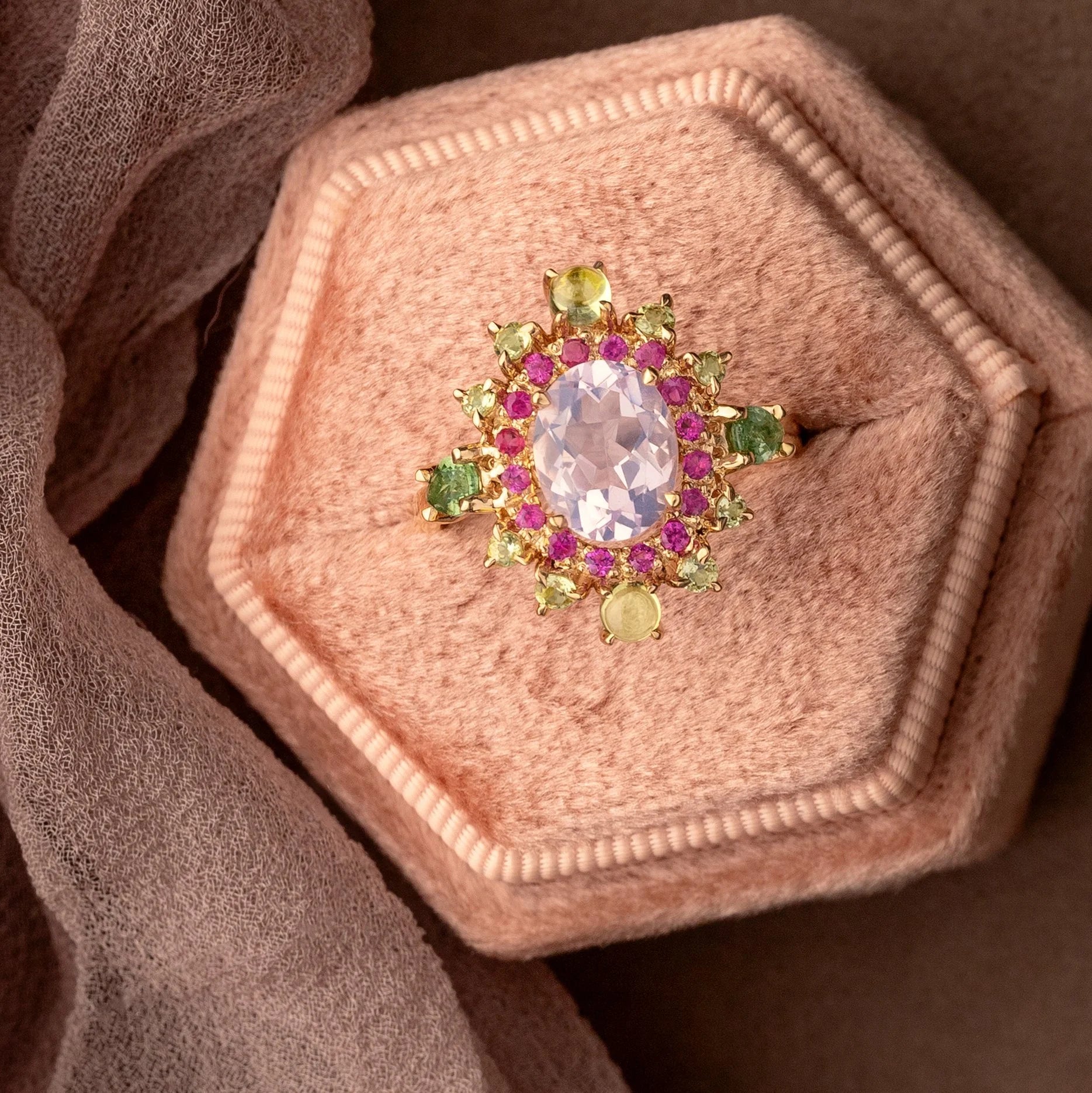 A gold ring with a large oval pale pink center stone and surrounding green and pink gemstones, displayed on a hexagonal blush pink velvet ring box with draped brown fabric.