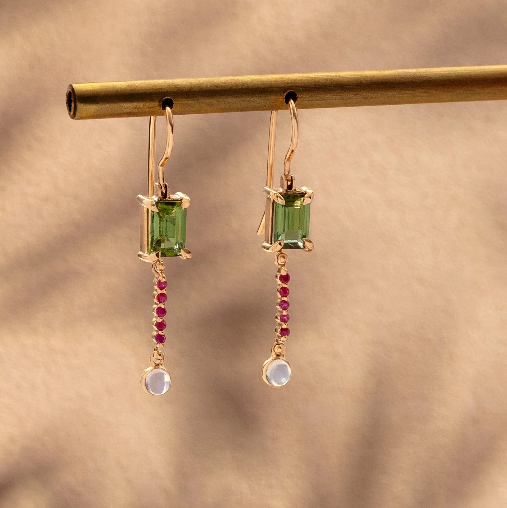 Pair of earrings with green gemstones and red rubies on a gold rod against a beige background