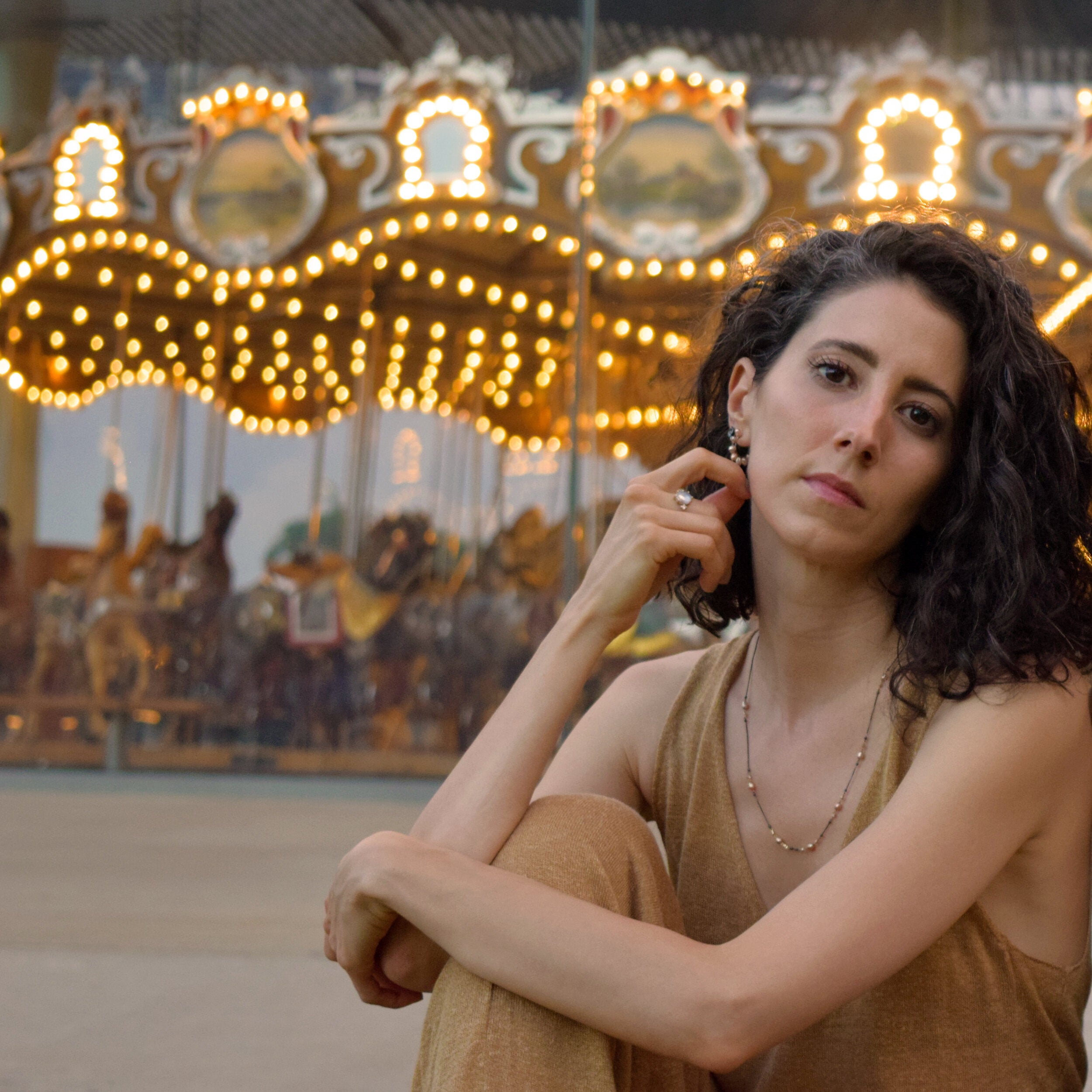 Woman sitting in front of a lit-up carousel at dusk.
