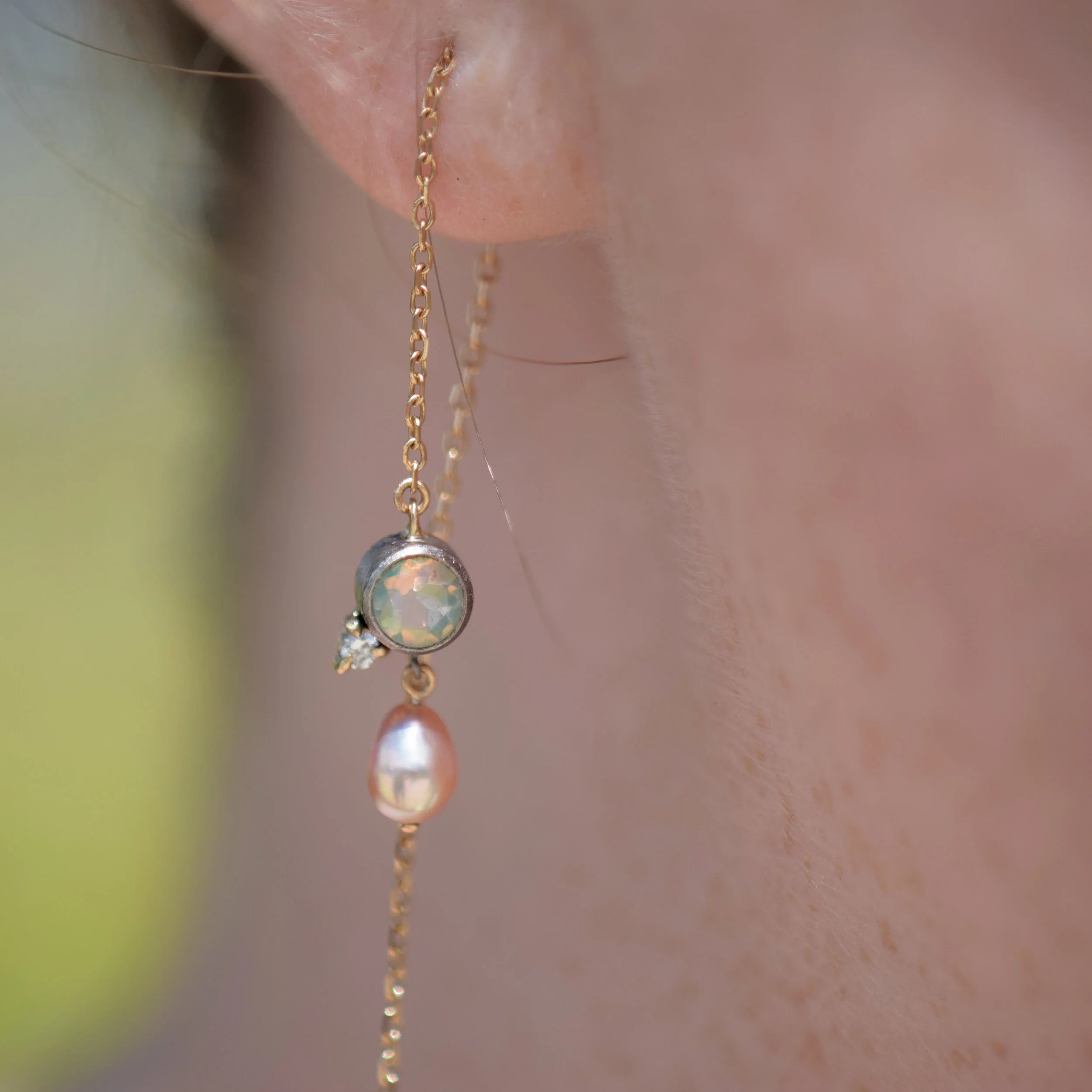 Delicate gold chain earring with a pearl and gemstone on skin against a blurred background